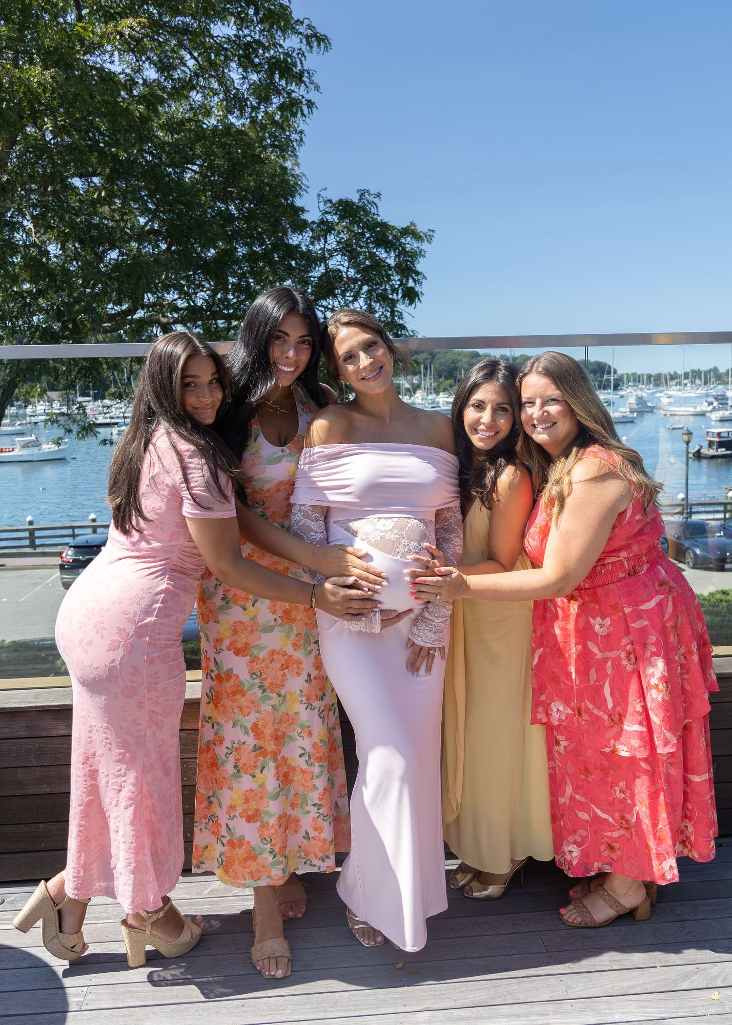 Pregnant woman in pink off-shoulder gown surrounded by four smiling friends on a sunny waterfront deck.