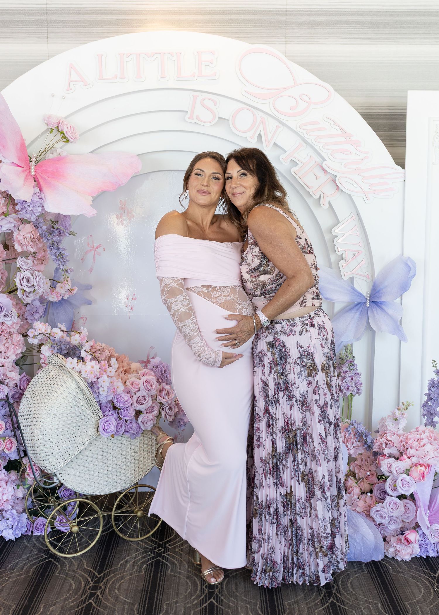 Pregnant woman in white off-shoulder gown posing with guest at floral butterfly baby shower backdrop.