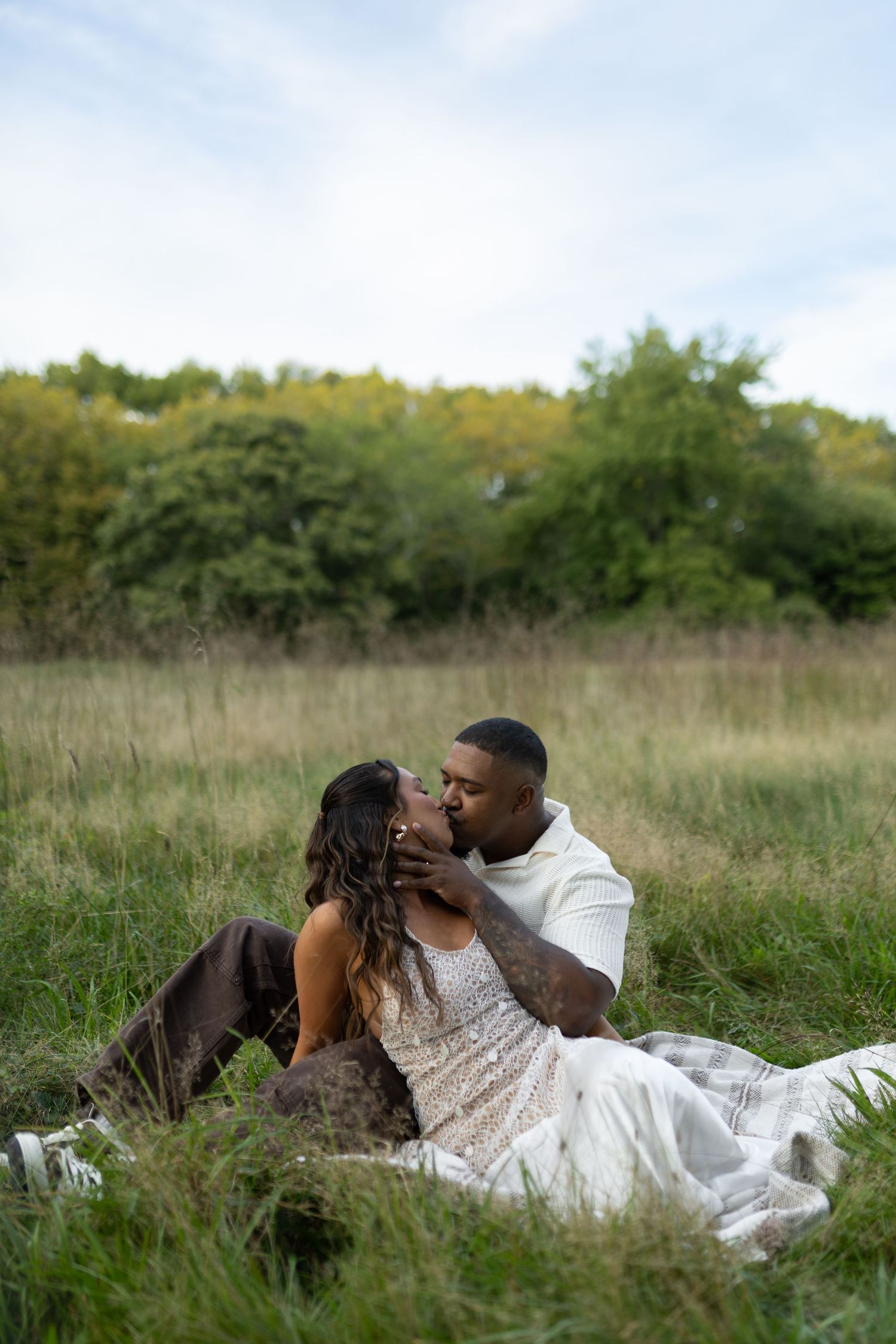 A couple shares a romantic kiss while sitting on a blanket in a lush green meadow with trees in the background.