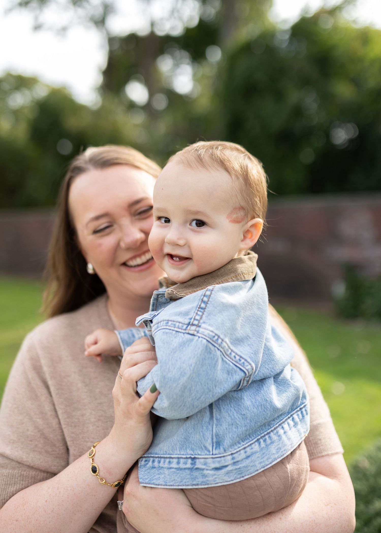 Smiling woman in a beige sweater holding a happy baby in a denim jacket outdoors in a sunny, leafy garden.