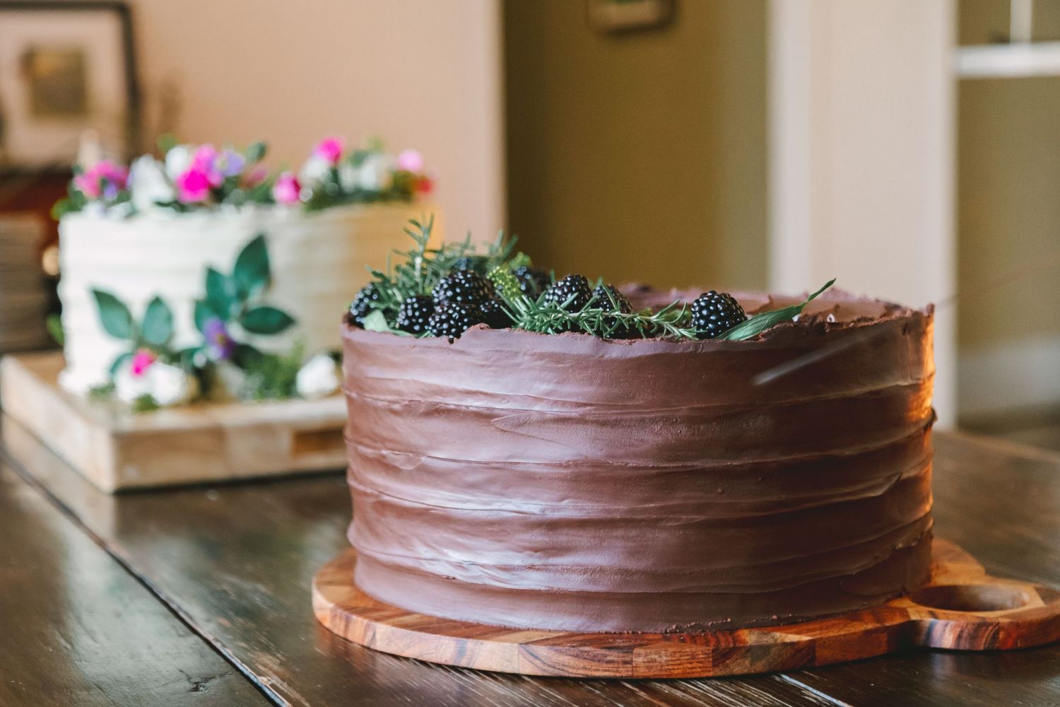 Rustic chocolate layer cake topped with blackberries and rosemary beside a white floral wedding cake.