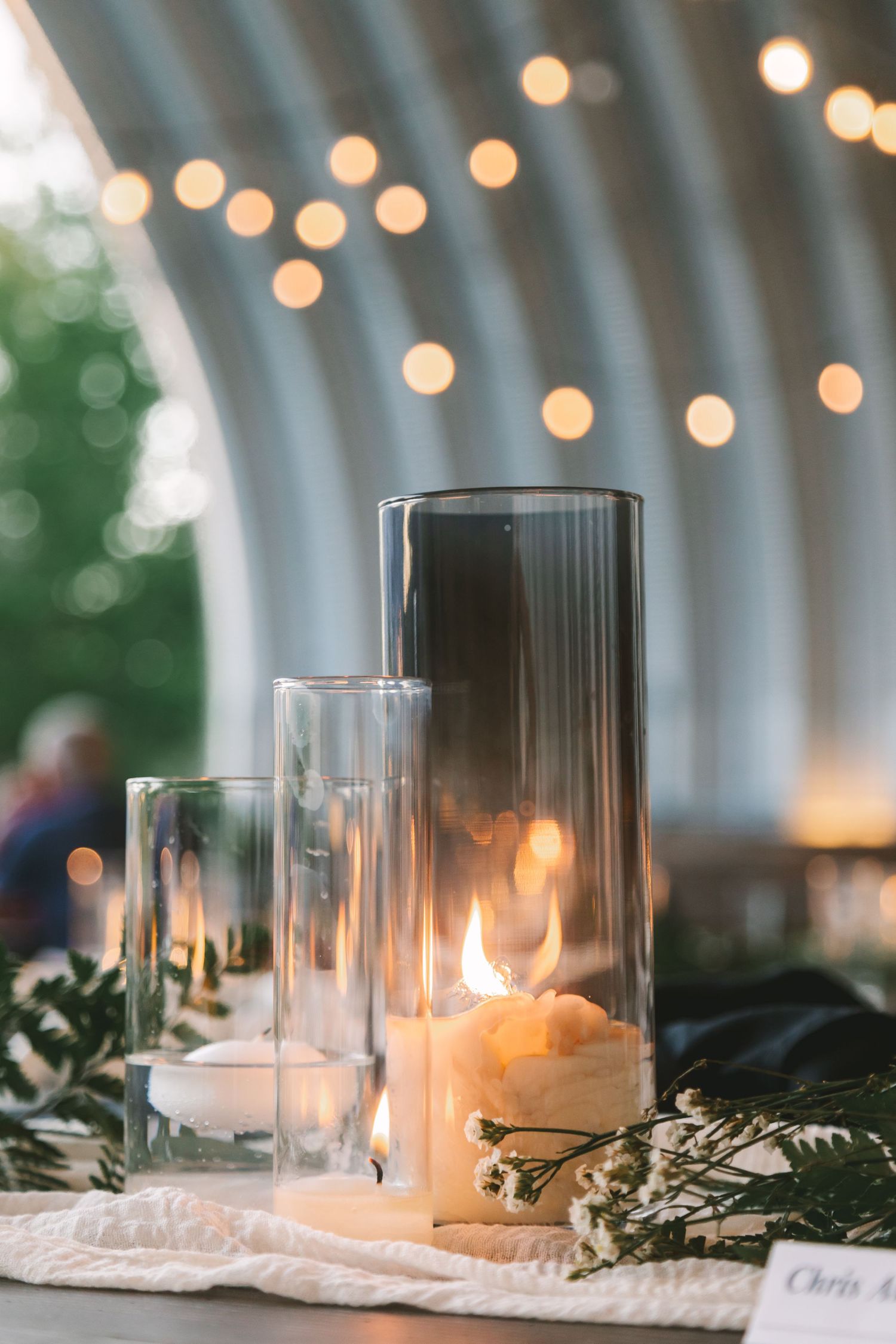 Elegant wedding reception table with lit candles in glass hurricanes, greenery, and warm string lights in the background.