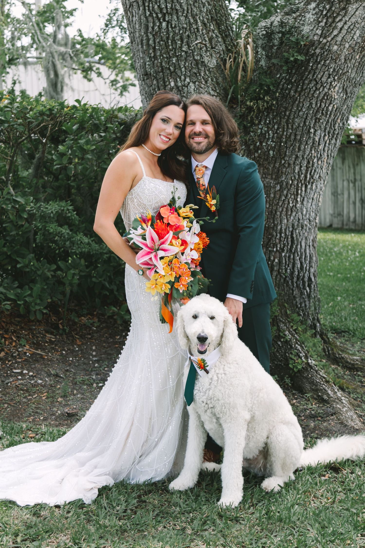 A bride and groom pose with their white Goldendoodle under a tree, the bride holding a vibrant tropical bouquet.