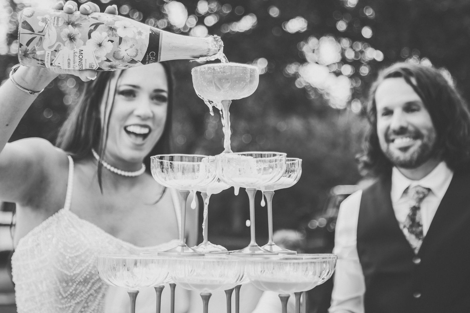 Black and white photo of laughing bride pouring champagne tower at wedding reception.