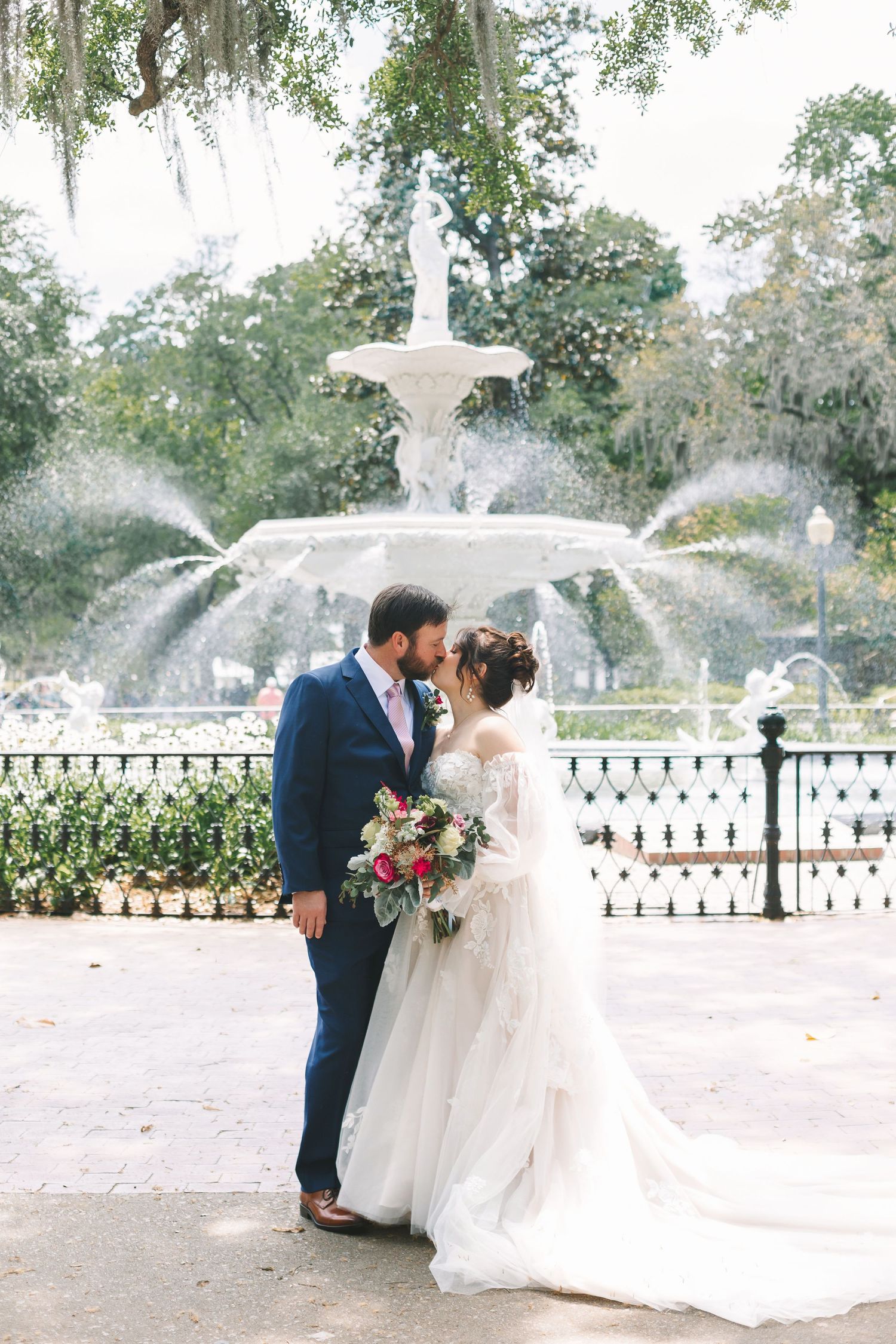 Bride and groom sharing a romantic kiss in front of a grand fountain in a lush garden setting.