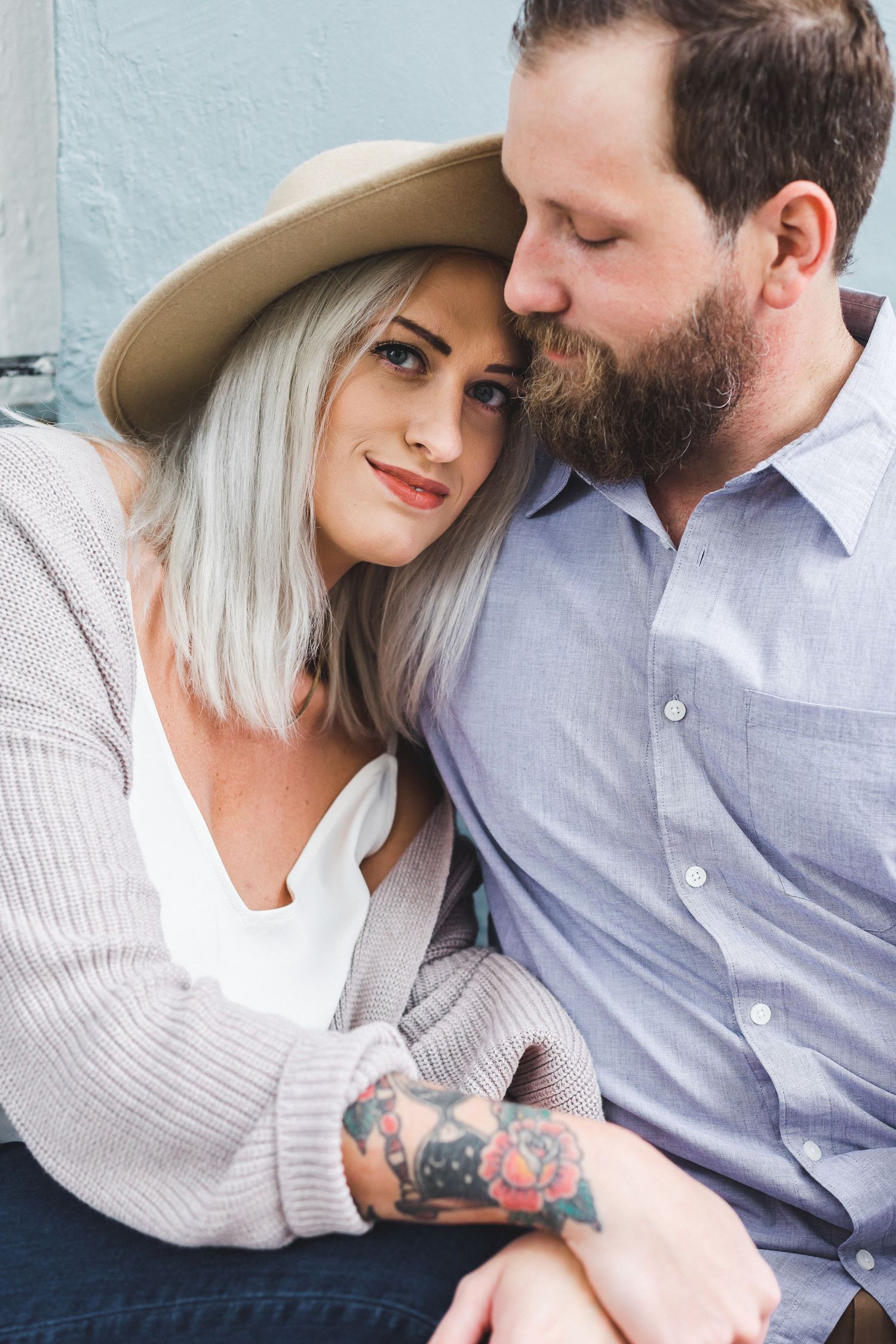 Tattooed woman in hat leans on bearded man's shoulder in cozy embrace.