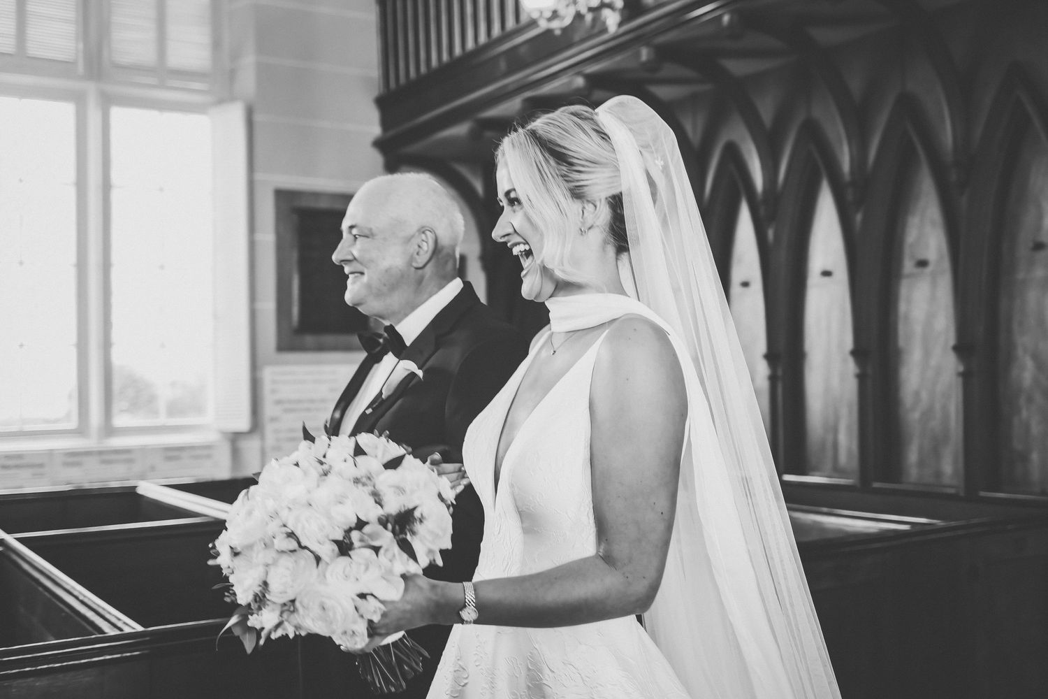 Black and white photo of smiling bride walking down church aisle with her father.