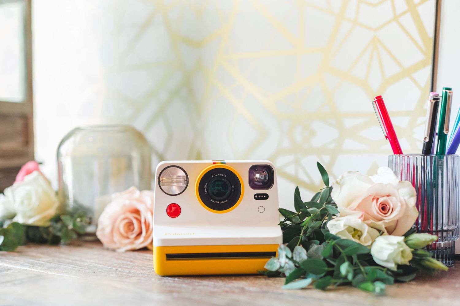 Yellow Polaroid Now instant camera surrounded by pink roses and greenery on a desk with pens and gold geometric wallpaper.