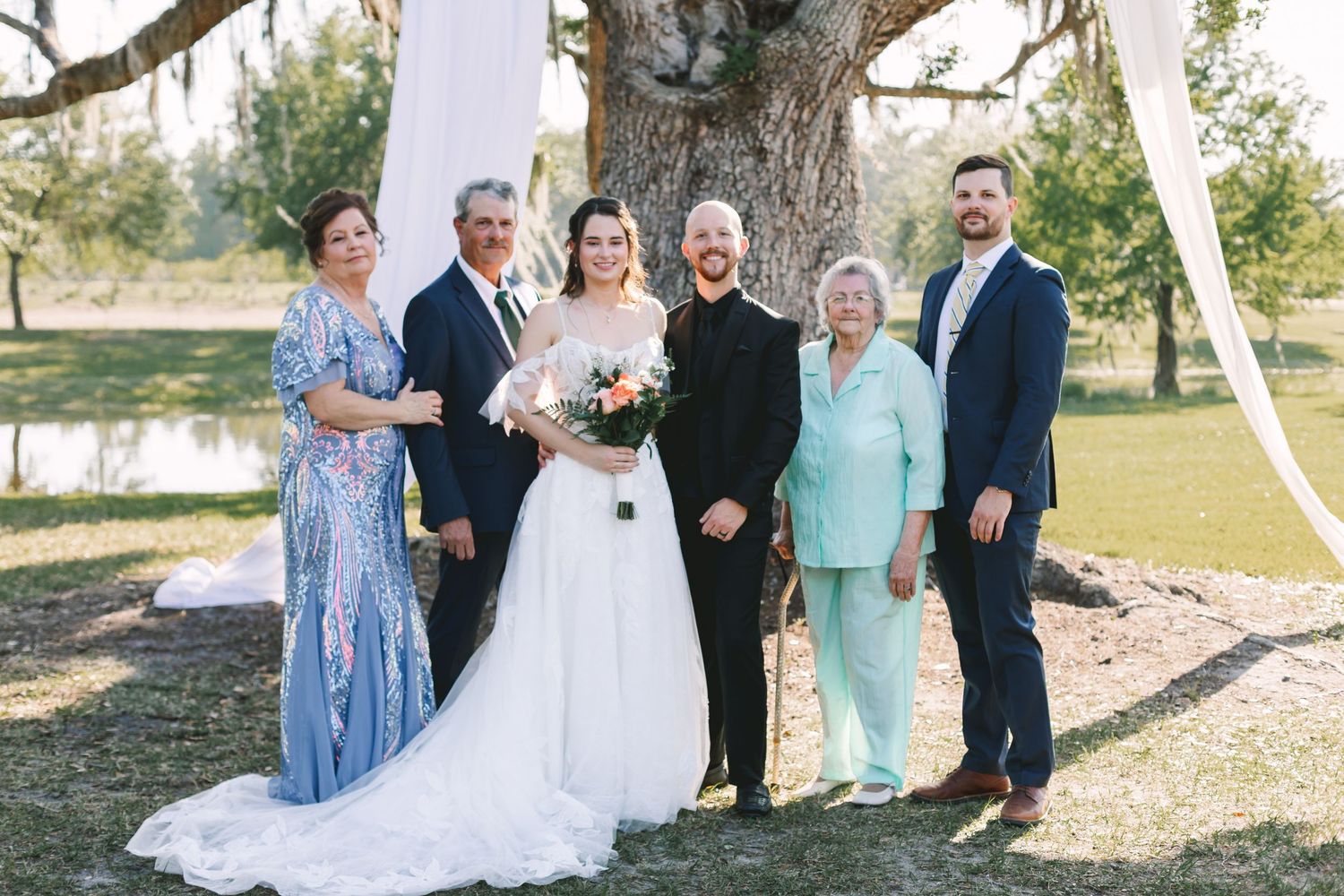 Wedding party family portrait with bride, groom, and guests posing under a large oak tree outdoors.