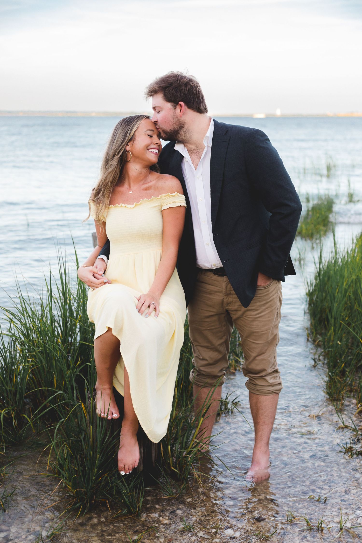 Man in blazer kissing woman in yellow dress while standing in shallow water.