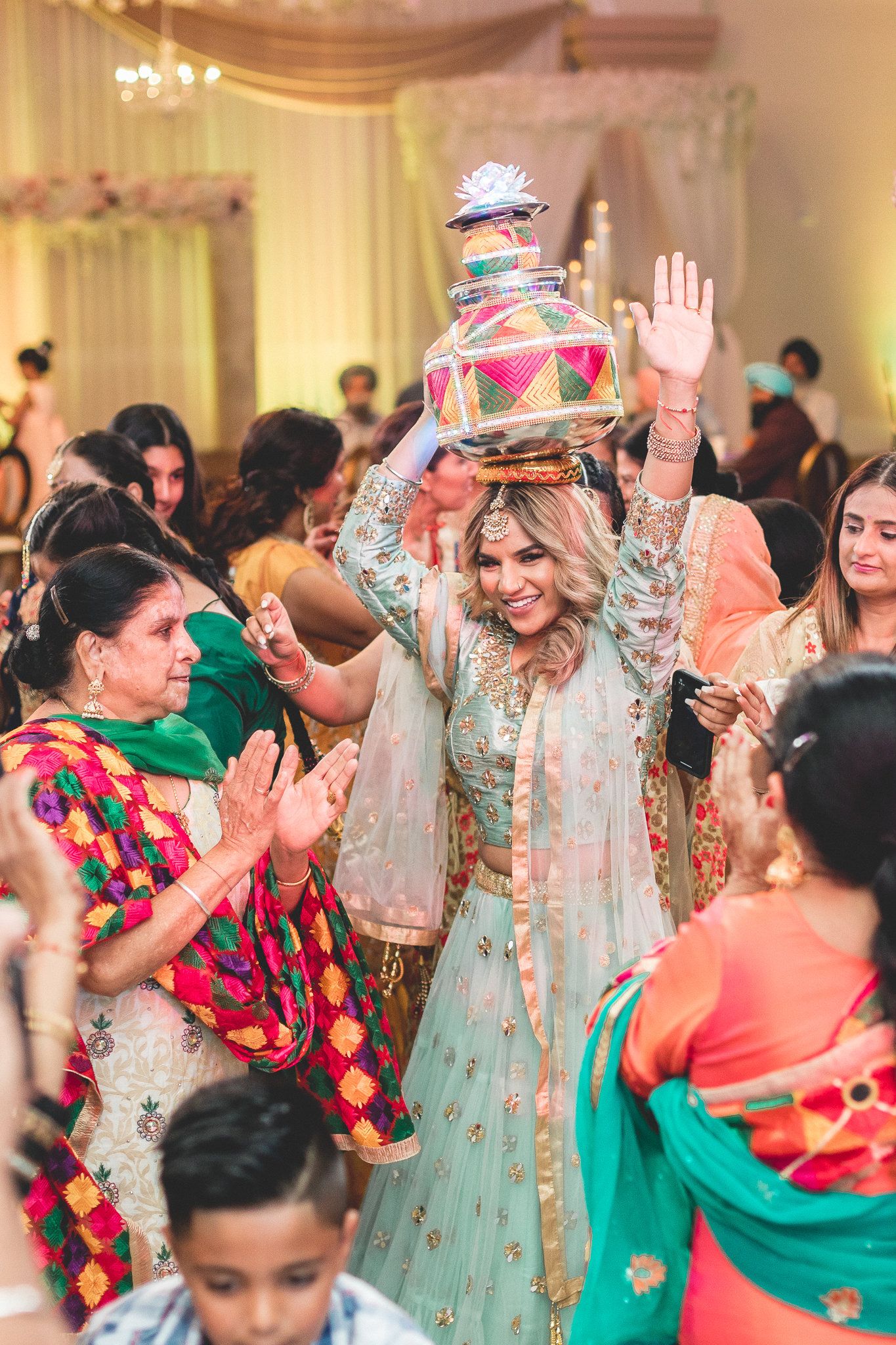 A joyful South Asian bride in mint lehenga balances a colorful pot on her head during a festive wedding ceremony.