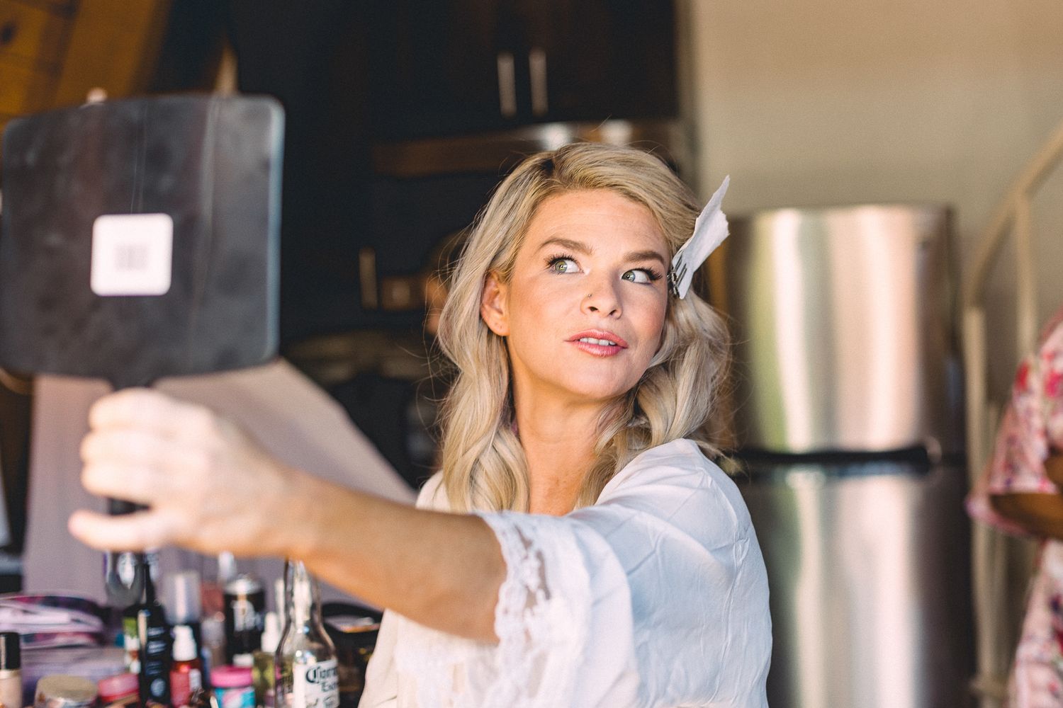 Blonde bride in white robe takes selfie with tablet mirror during wedding preparations.