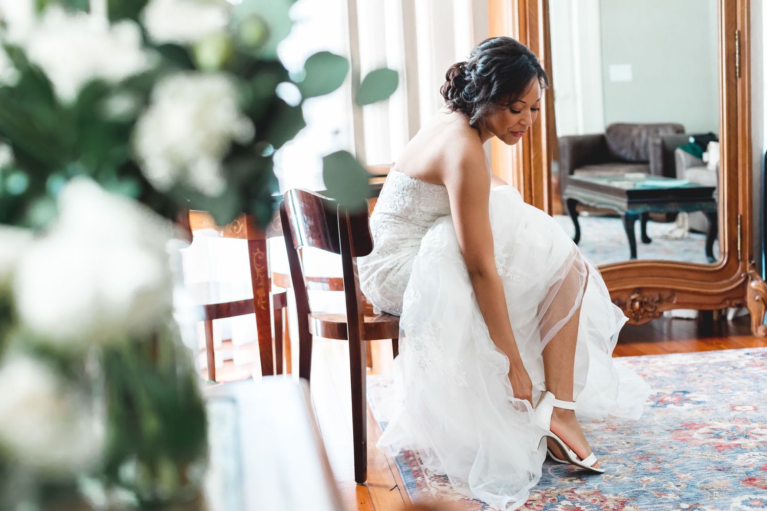 Bride in a strapless white wedding gown putting on heels while seated near a vintage mirror indoors.