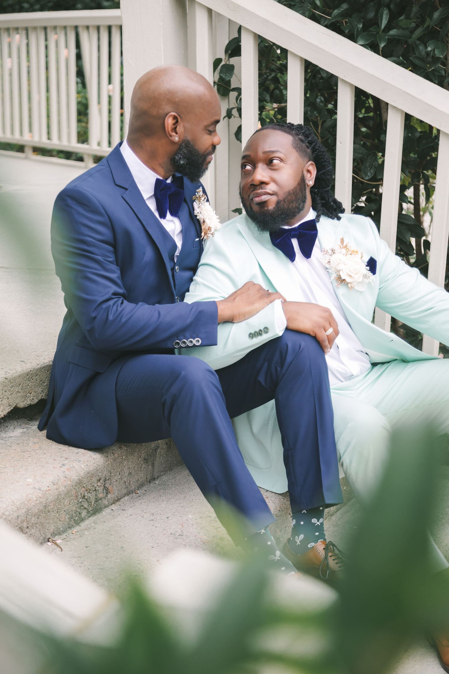 Two grooms in blue and mint suits sit on outdoor steps sharing a loving glance.