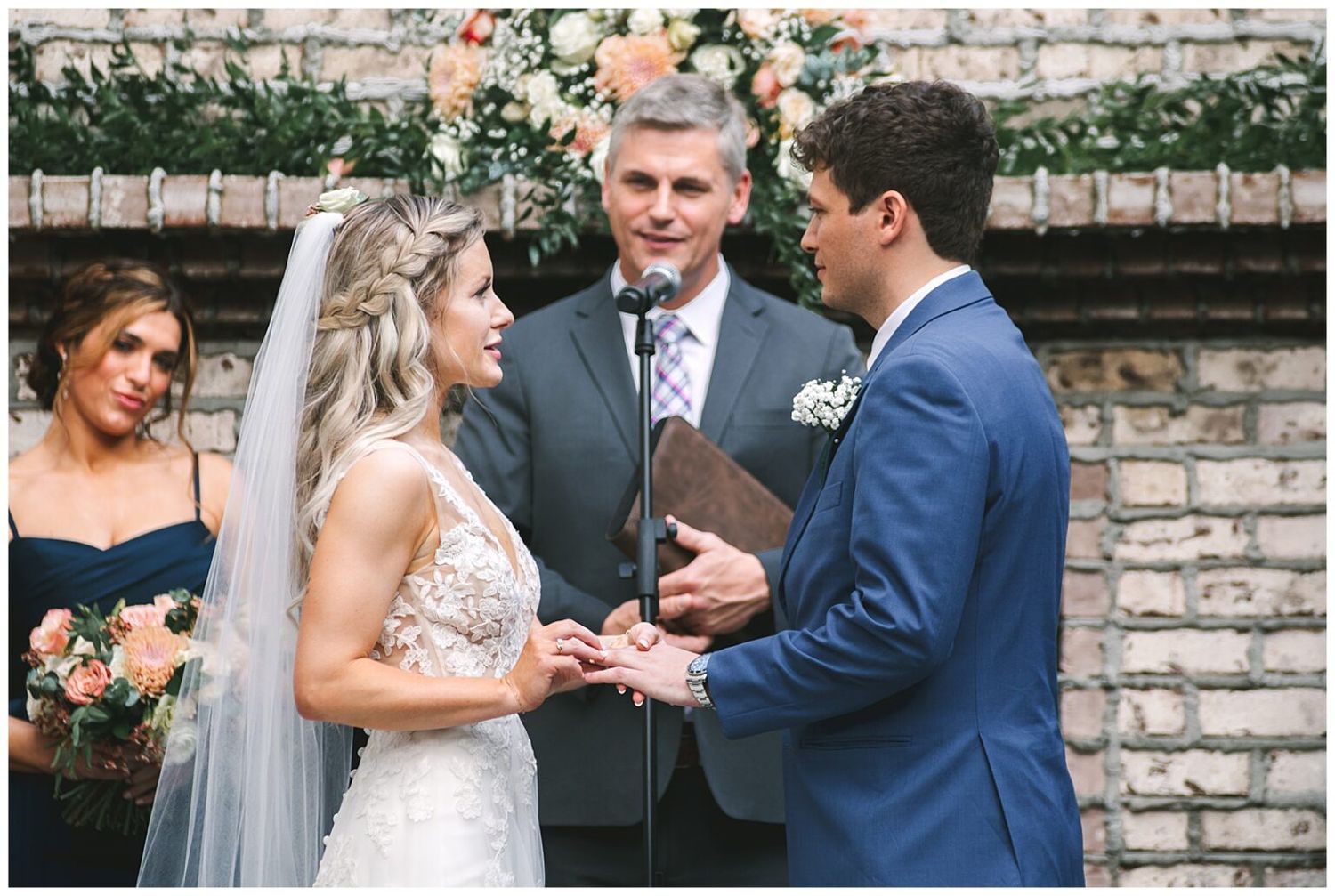Groom reads vows to bride during outdoor wedding ceremony at a rustic brick venue with floral arch backdrop.