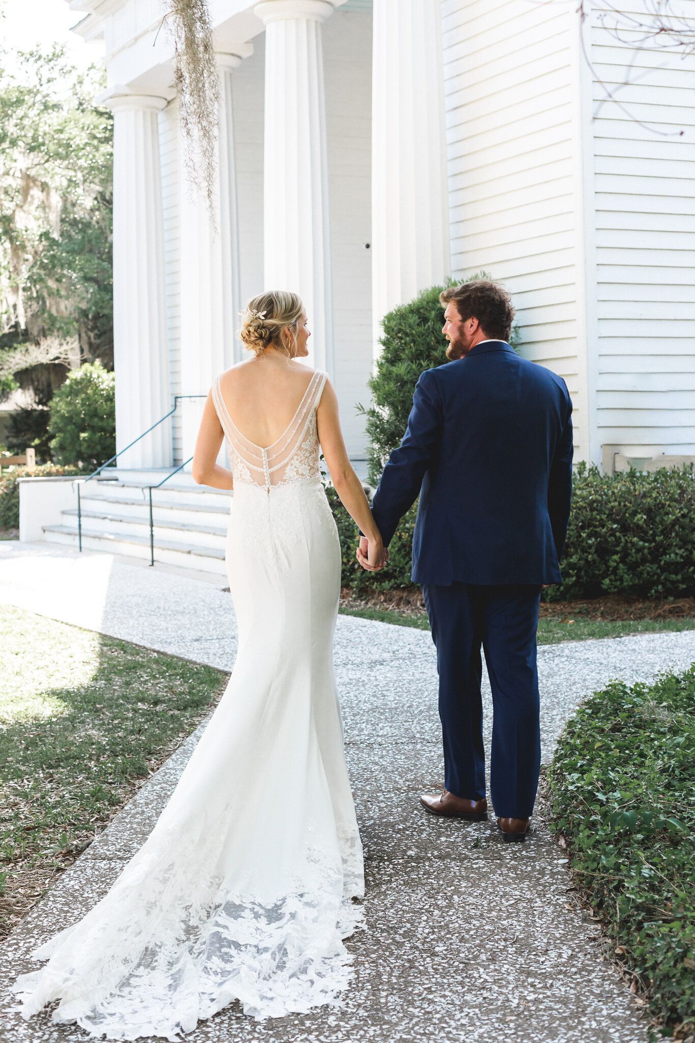 Bride in backless lace wedding gown holding hands with groom walking toward white church.