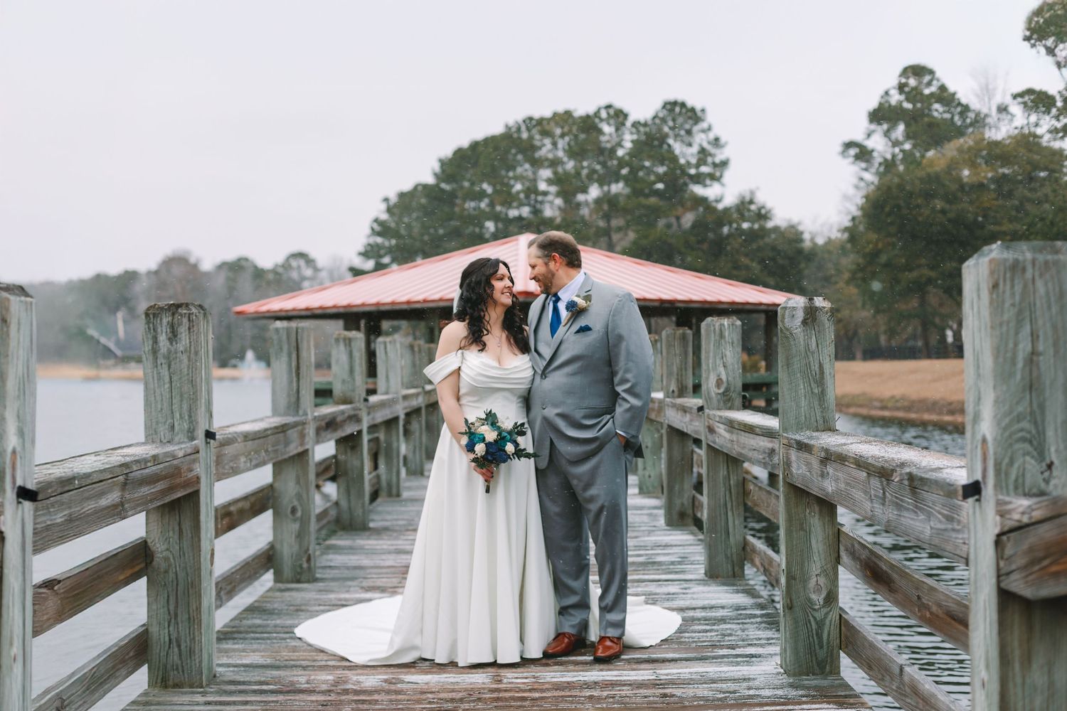 Bride and groom share romantic moment on wooden dock by water.