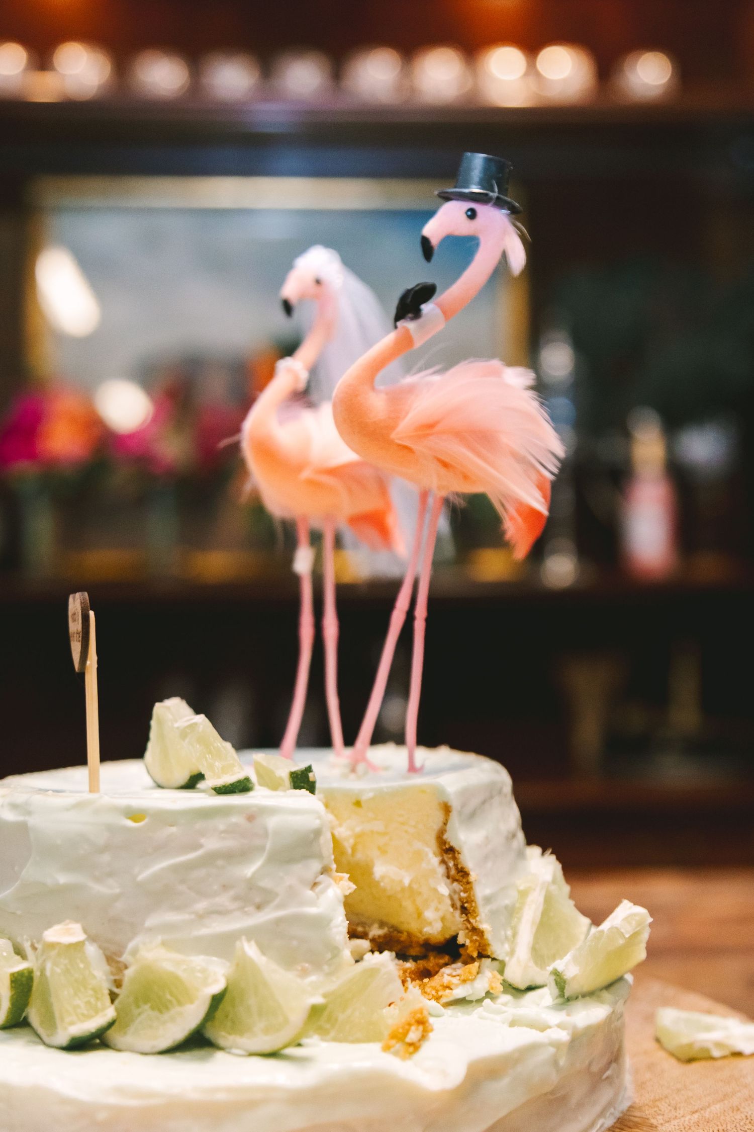 A white wedding cake topped with pink flamingo cake toppers, decorated with lime slices, with a slice removed.