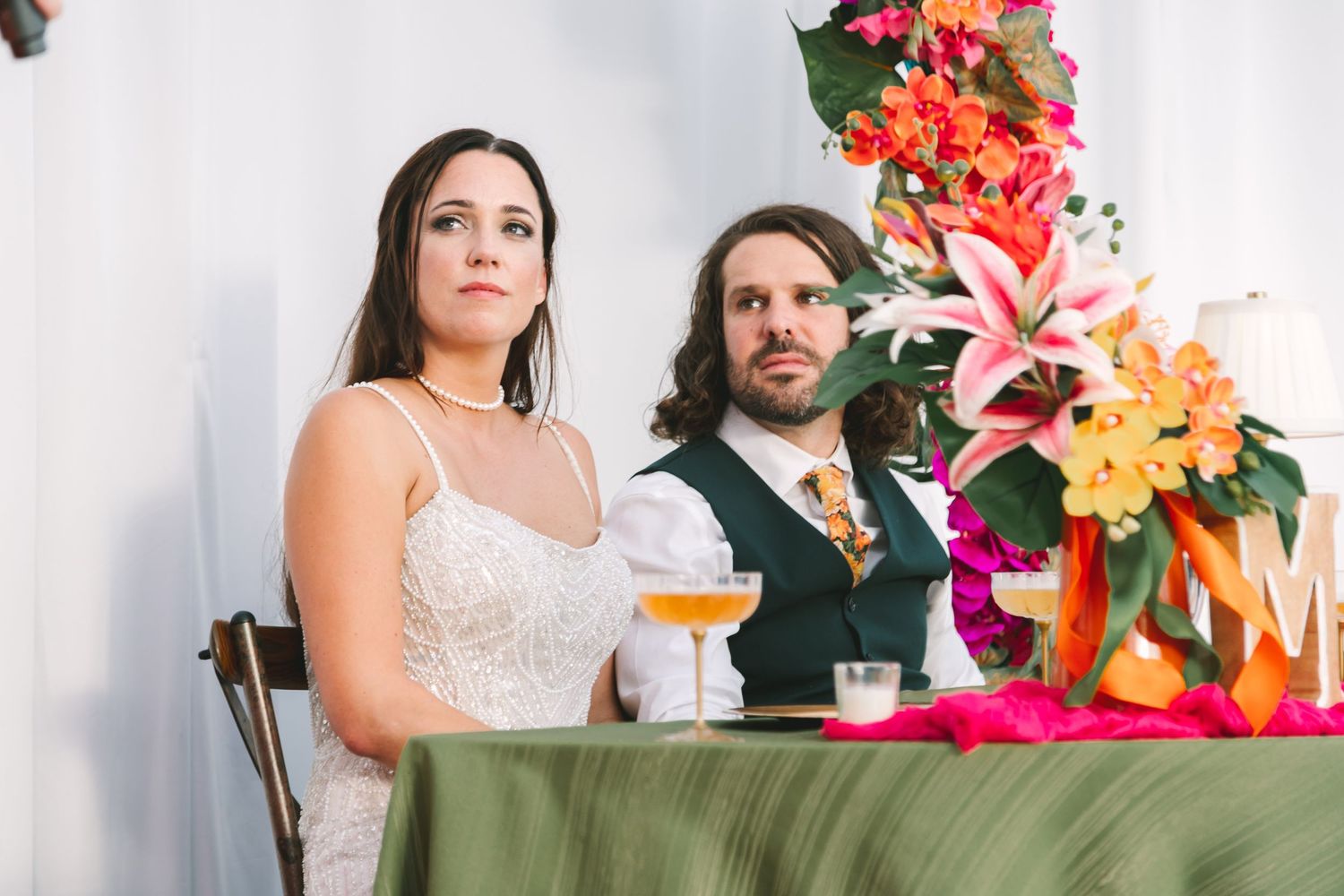 Bride and groom seated at sweetheart table with tropical floral arrangements at wedding reception.