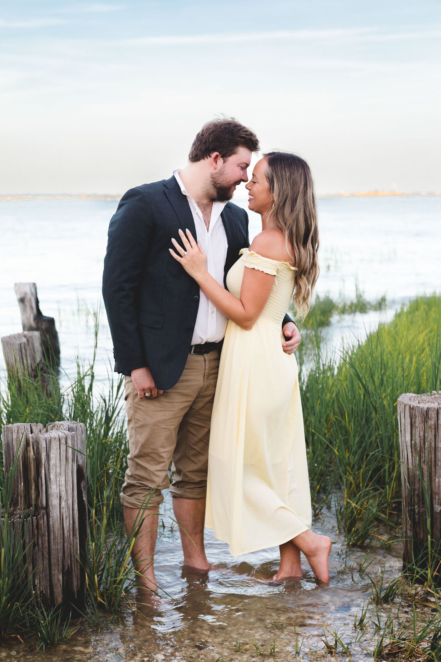 Romantic couple nearly kissing while standing barefoot in shallow water at golden hour.