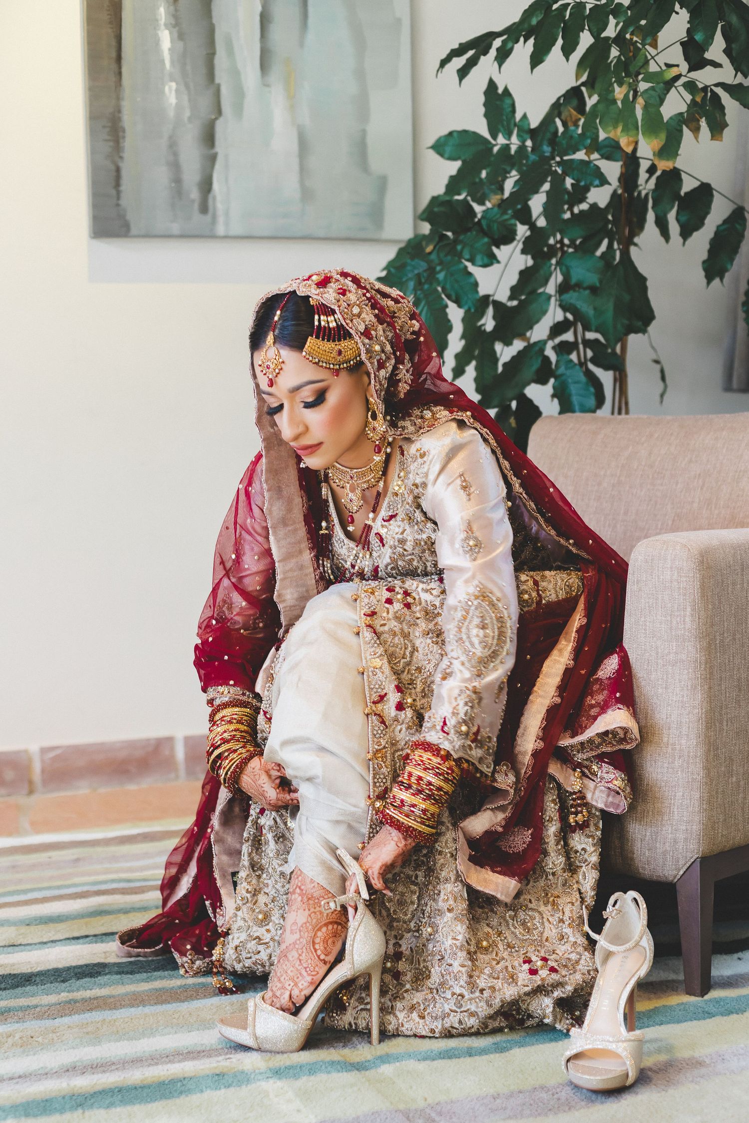 South Asian bride in ivory and red bridal lehenga with gold jewelry putting on silver heels.