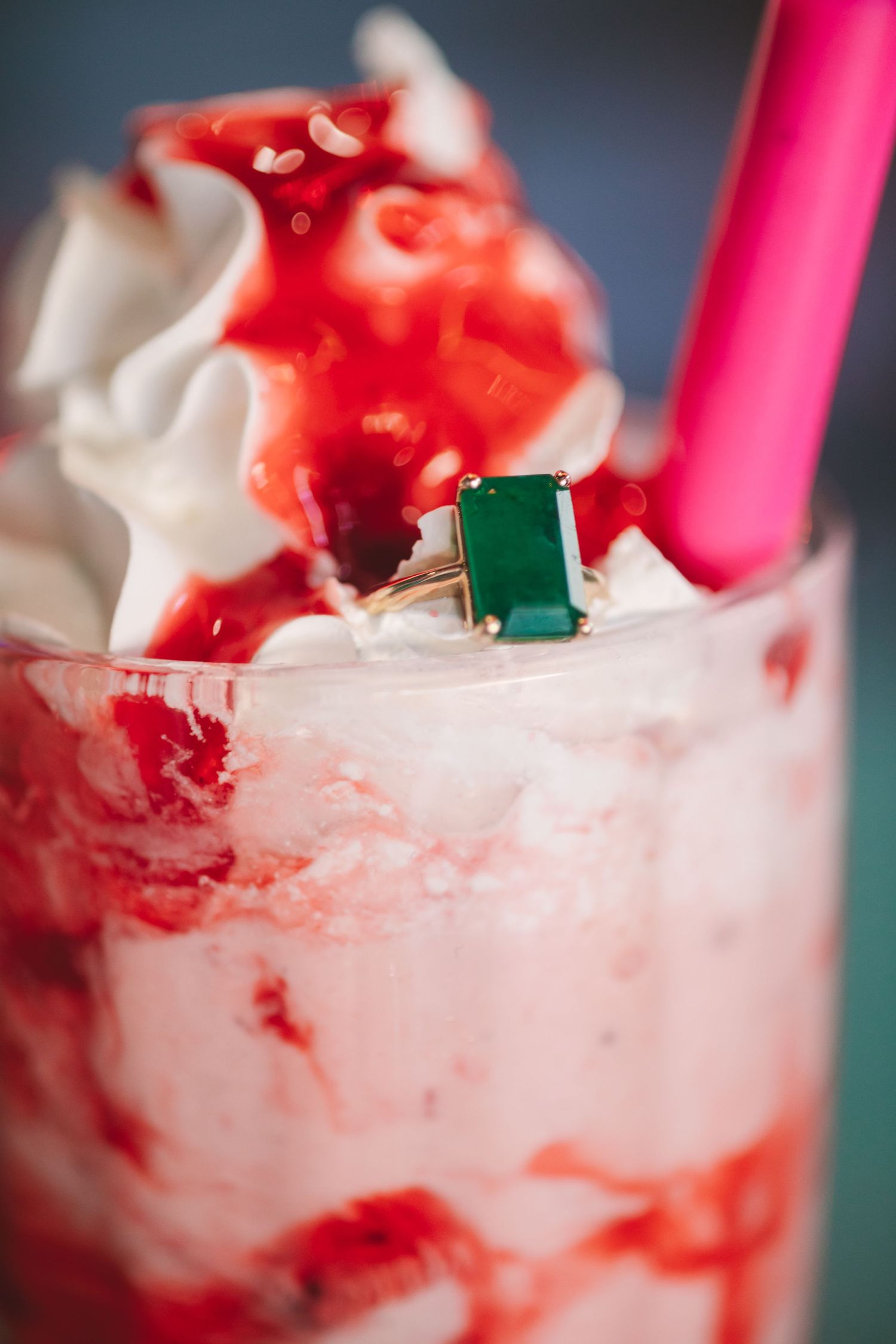 Gold ring with emerald green gemstone resting on a strawberry milkshake with whipped cream and pink straw.