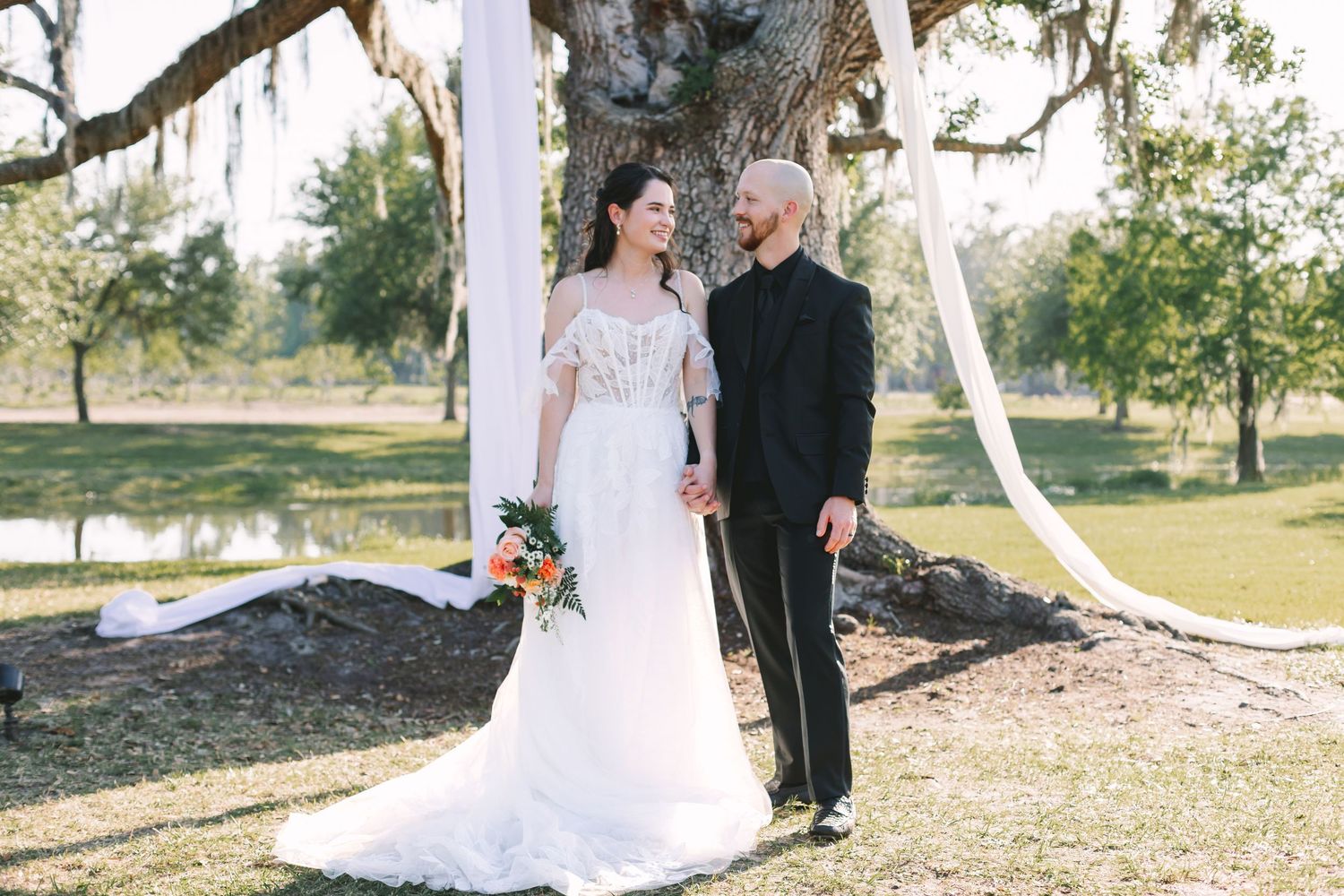 Bride and groom pose together under draped white fabric near large oak tree during outdoor wedding ceremony.