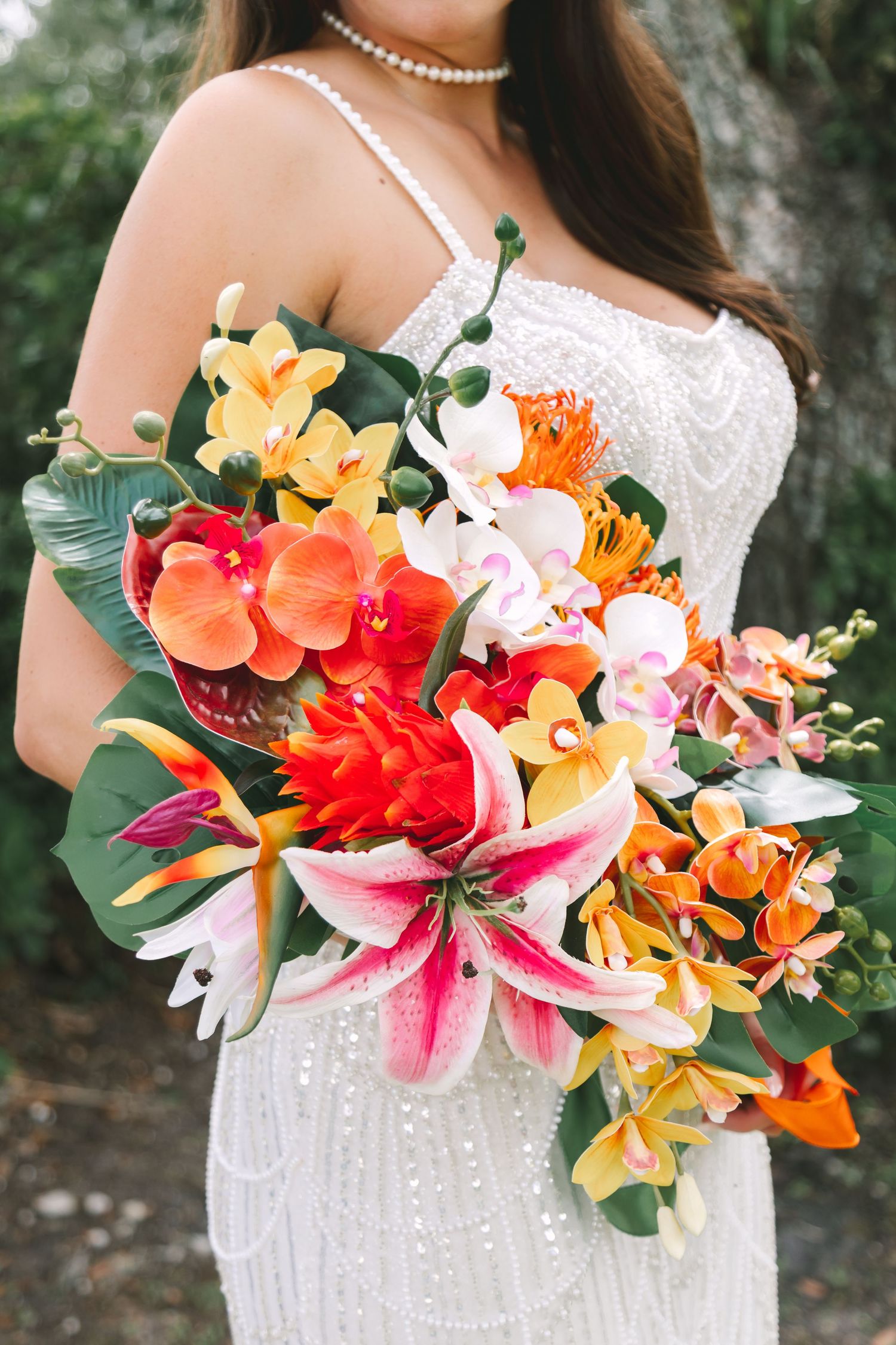Bride holds vibrant tropical bouquet with stargazer lilies, orchids, and orange flowers in white beaded gown.