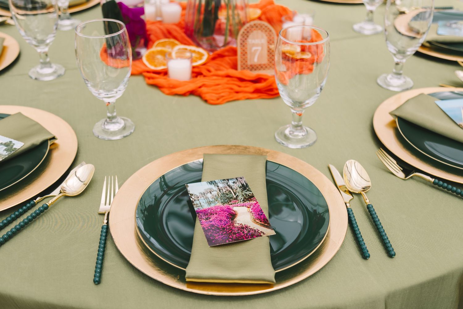 Elegant wedding table setting with green tablecloth, gold chargers, dark plates, and colorful centerpiece.
