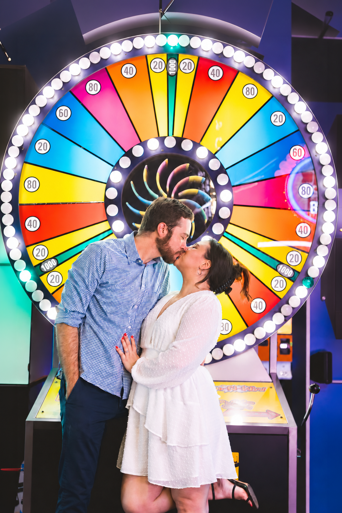 Couple kissing romantically in front of a colorful spinning prize wheel at an arcade.
