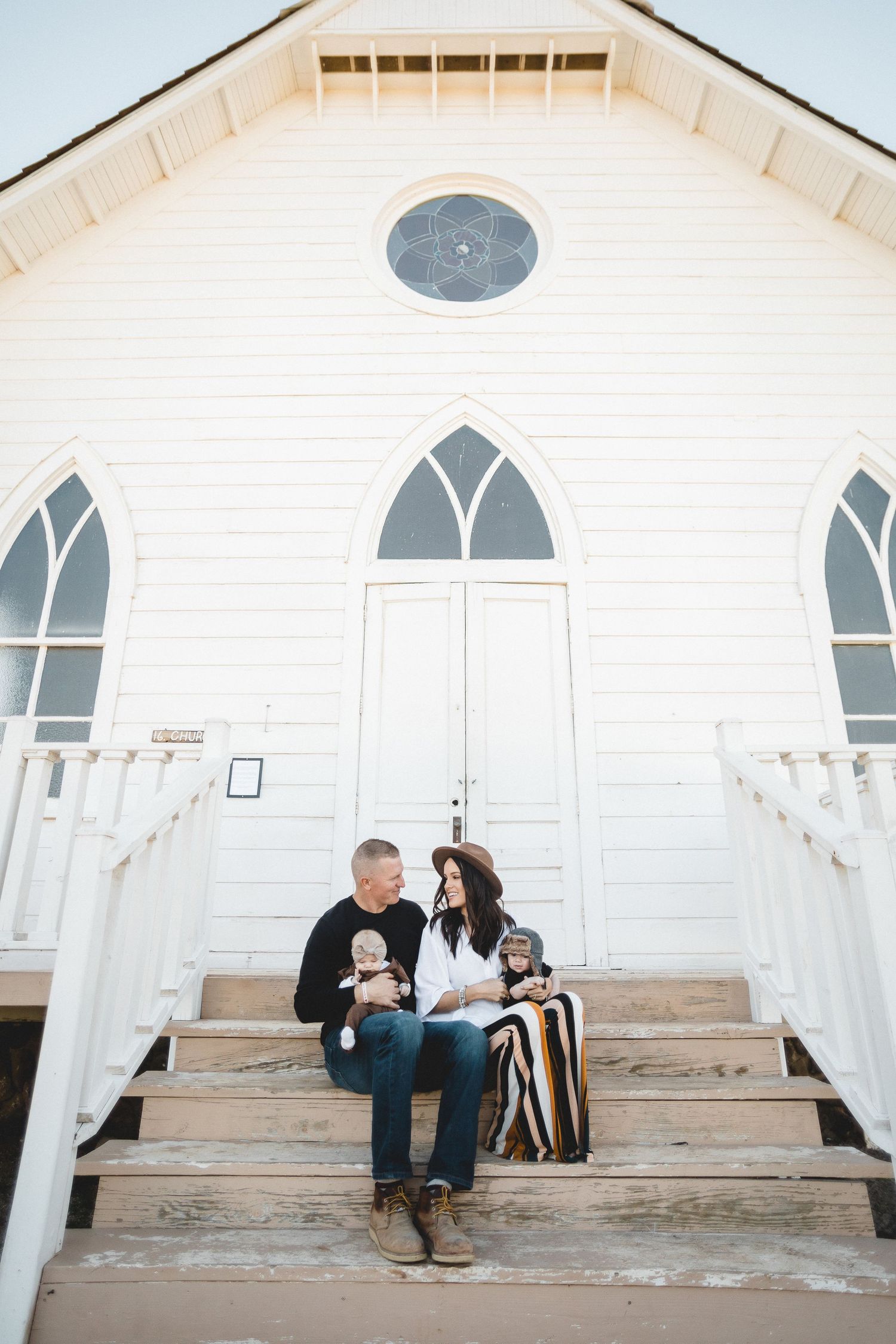 Family sitting on church steps with two babies, white chapel with gothic windows backdrop.