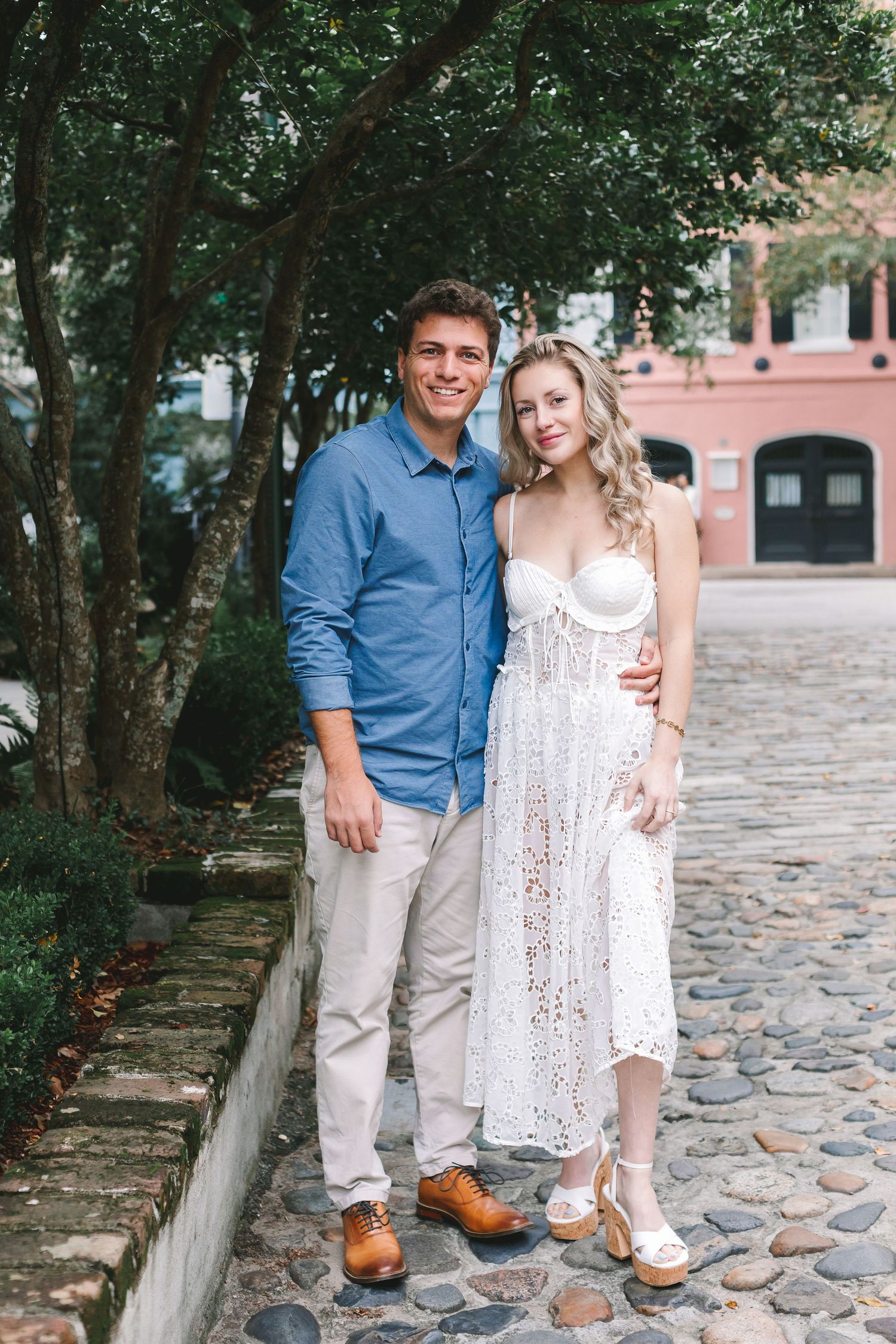 Smiling couple posing together on a charming cobblestone path surrounded by lush green trees.