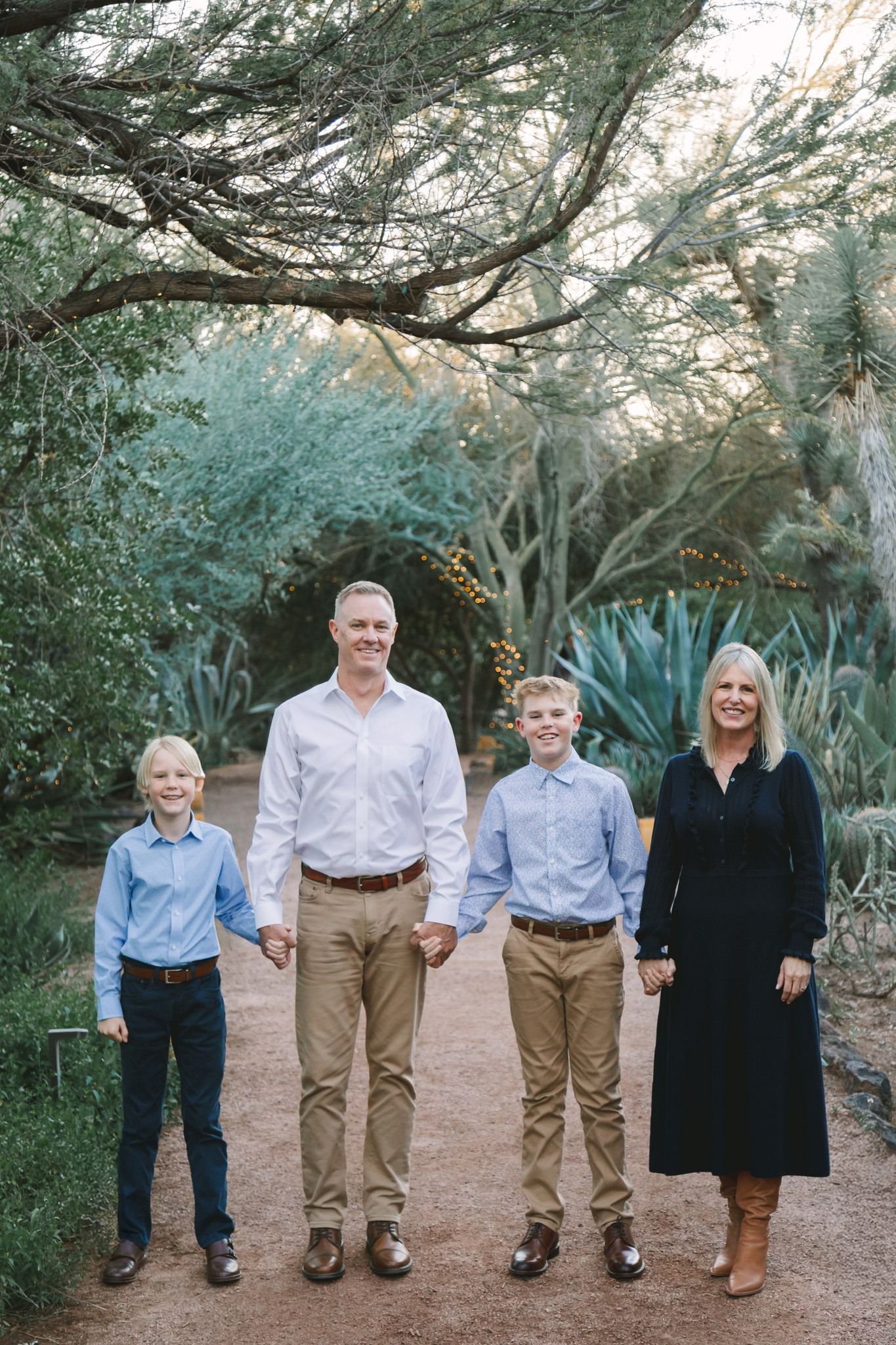 Family of four holding hands on a desert path surrounded by cacti and lush greenery.