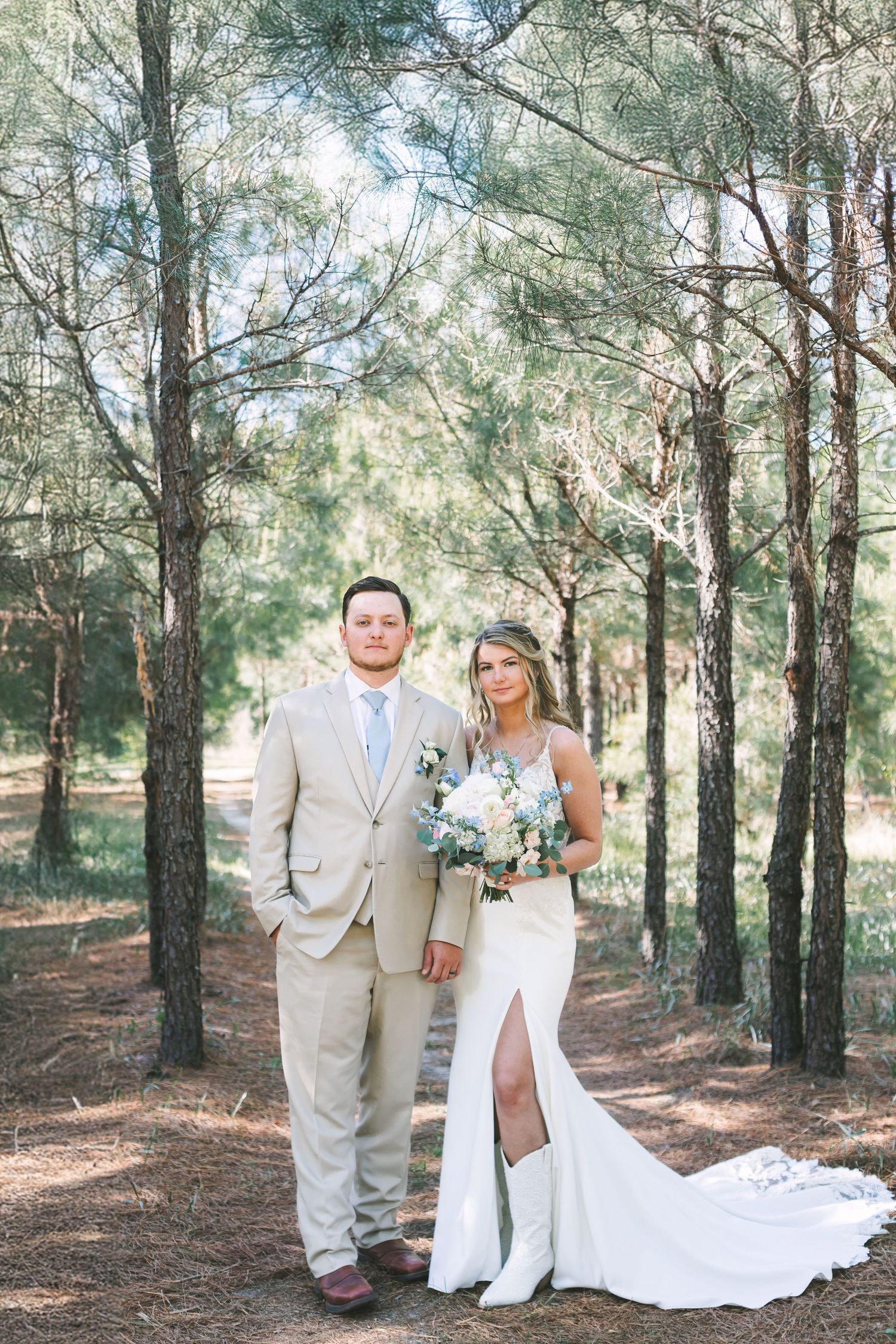 Bride and groom posing together in a pine forest, bride holding white bouquet in fitted wedding gown.