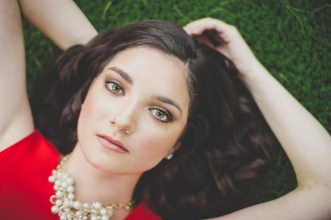 Beautiful brunette woman in red dress lying on grass, wearing pearl necklace, looking up.