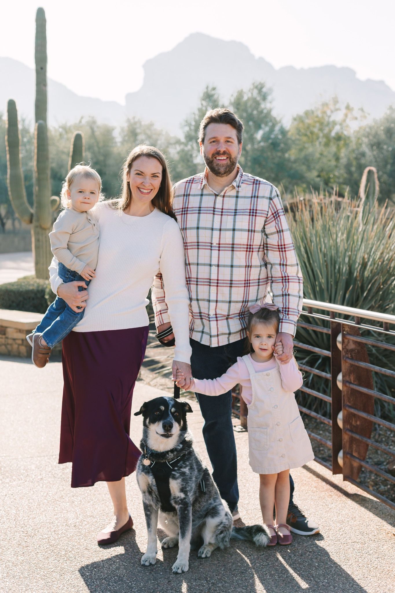 Family of four with a dog posing outdoors near cacti and desert mountains in warm sunlight.