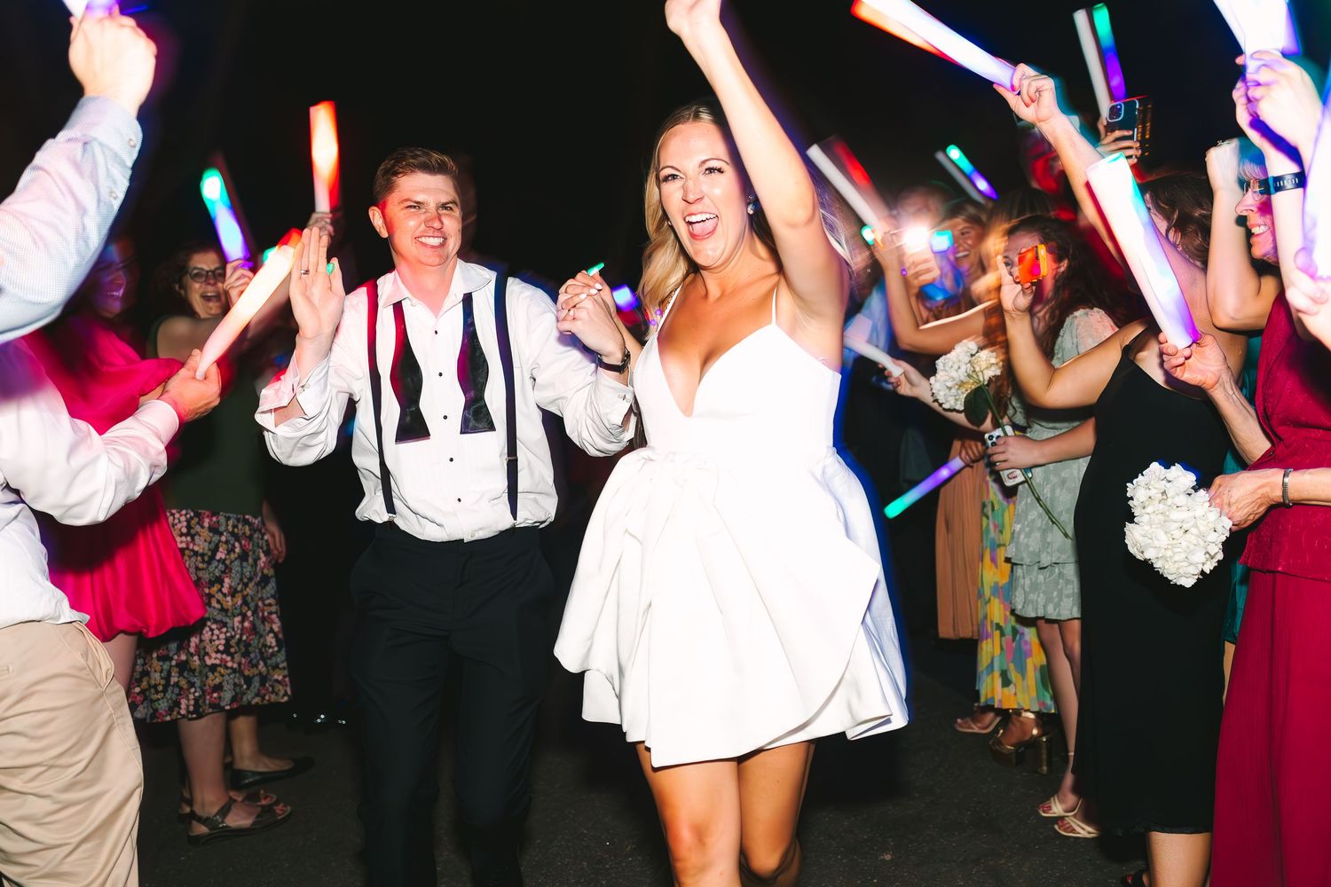 Bride and groom exit wedding reception through tunnel of glowing light sticks held by guests.