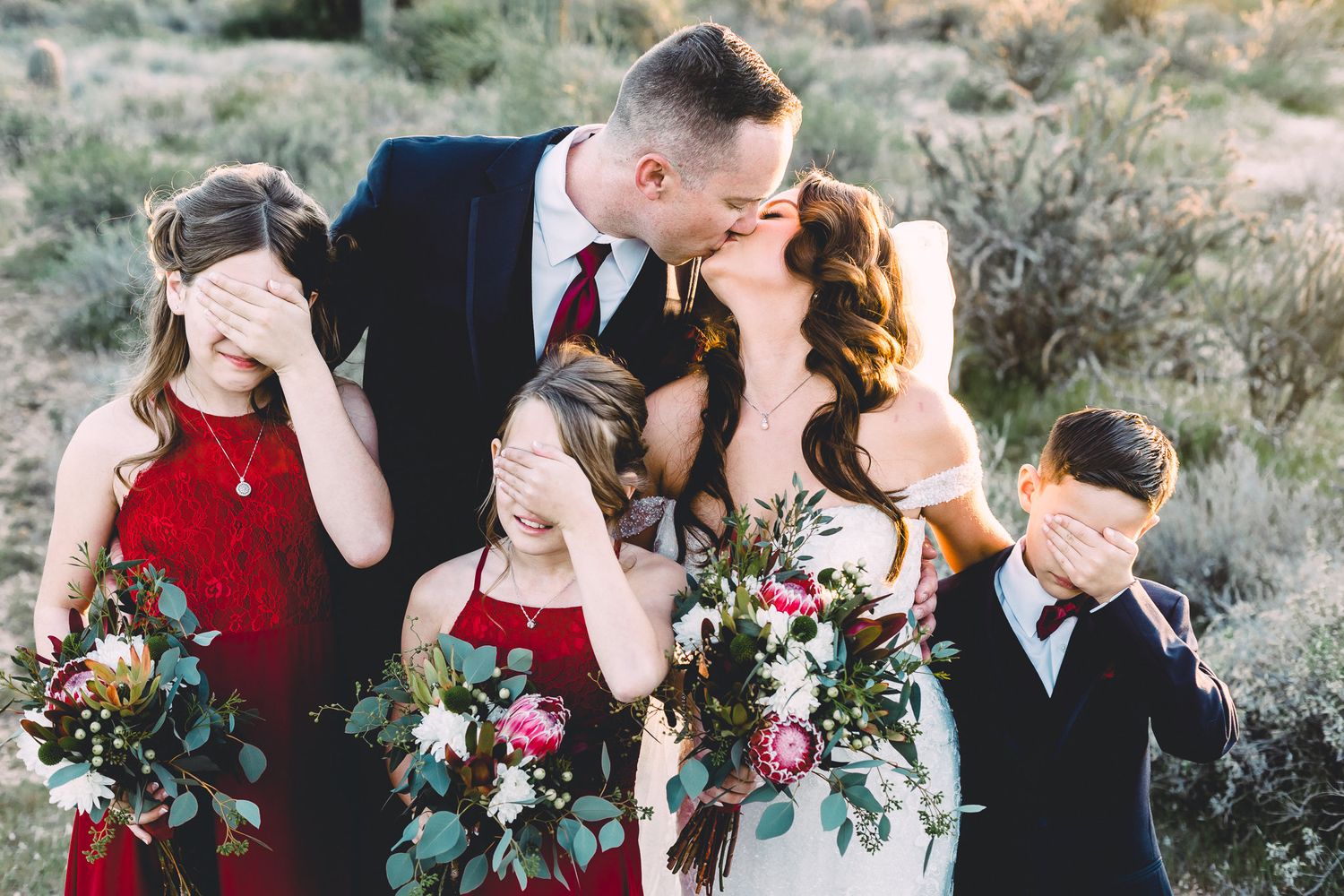 Bride and groom kissing while children playfully cover their eyes at outdoor wedding.