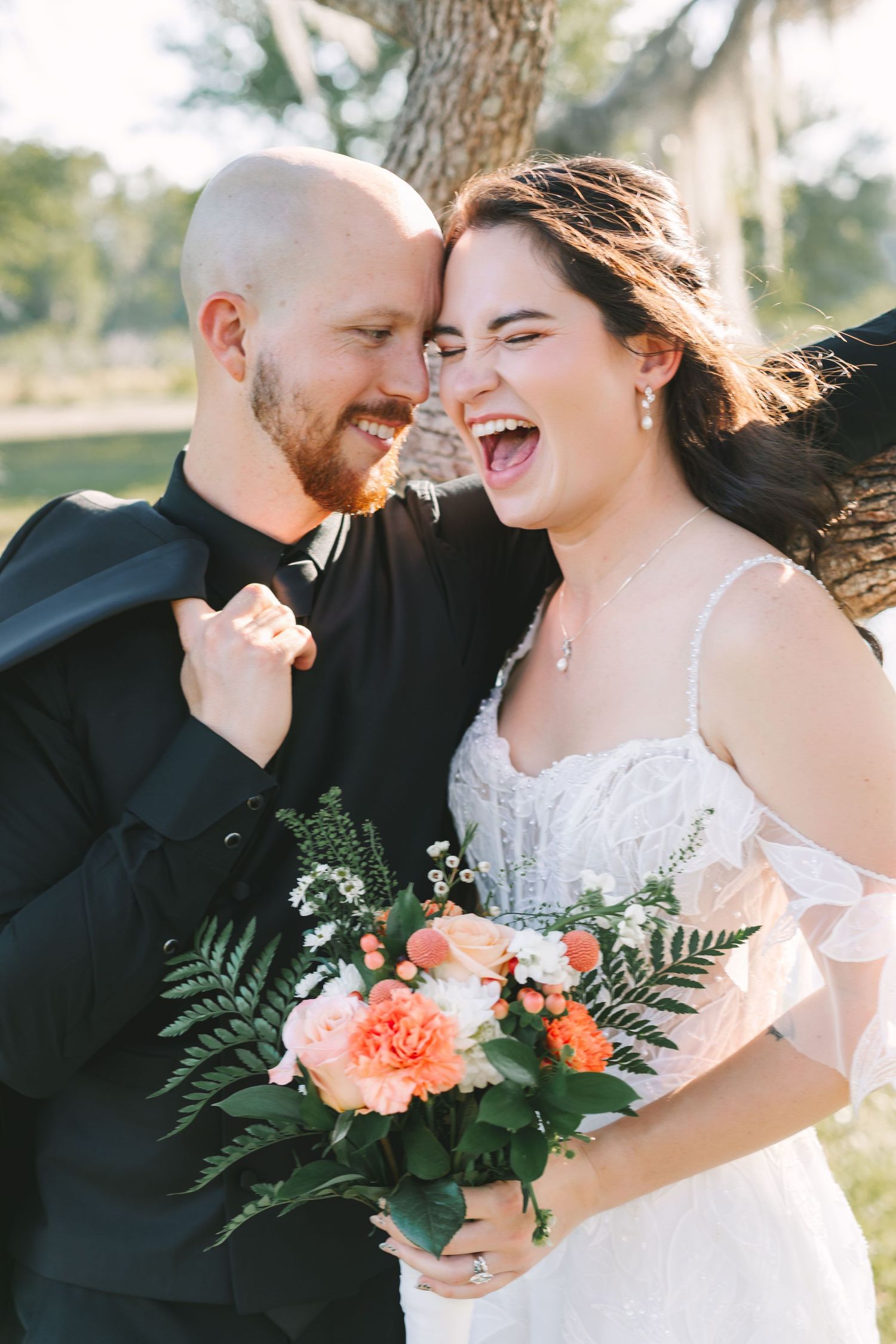 Joyful bride laughing with groom nuzzling her cheek, holding coral bridal bouquet at outdoor Florida wedding.