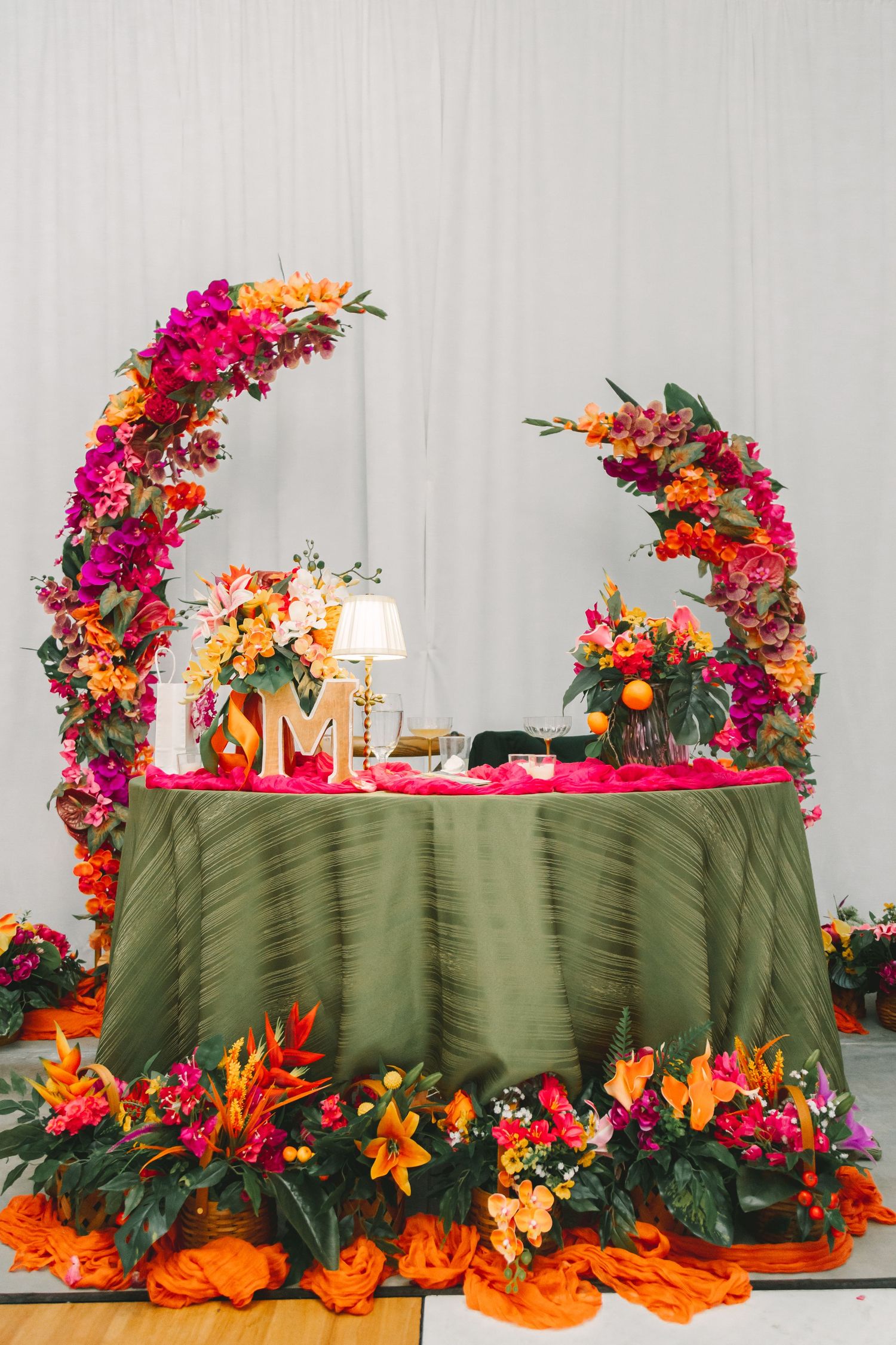 Sweetheart table decorated with vibrant orange and pink floral arches and green tablecloth at wedding reception.