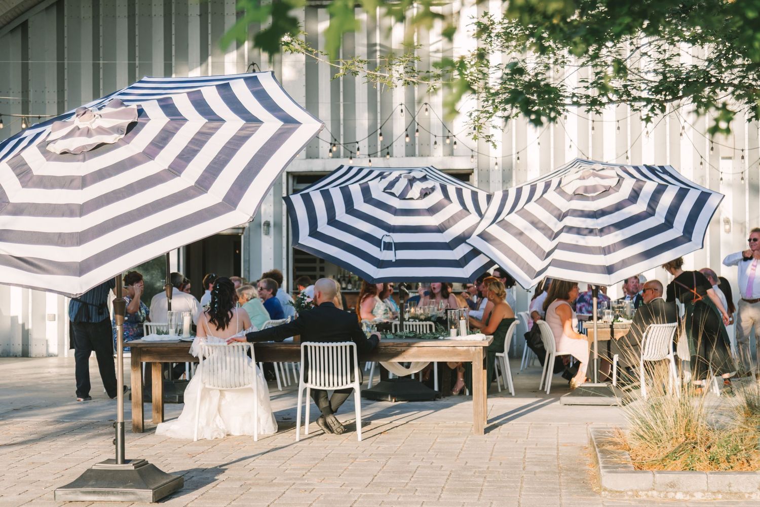 Outdoor dining area with blue and white striped umbrellas, guests seated at wooden tables on a sunny patio.