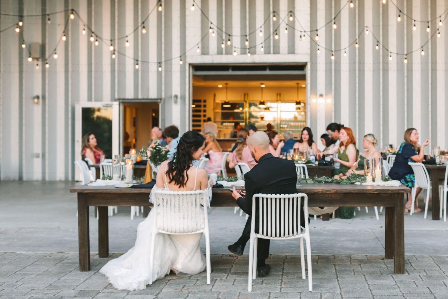 Wedding reception guests dine at rustic wooden tables under string lights in a modern barn venue.