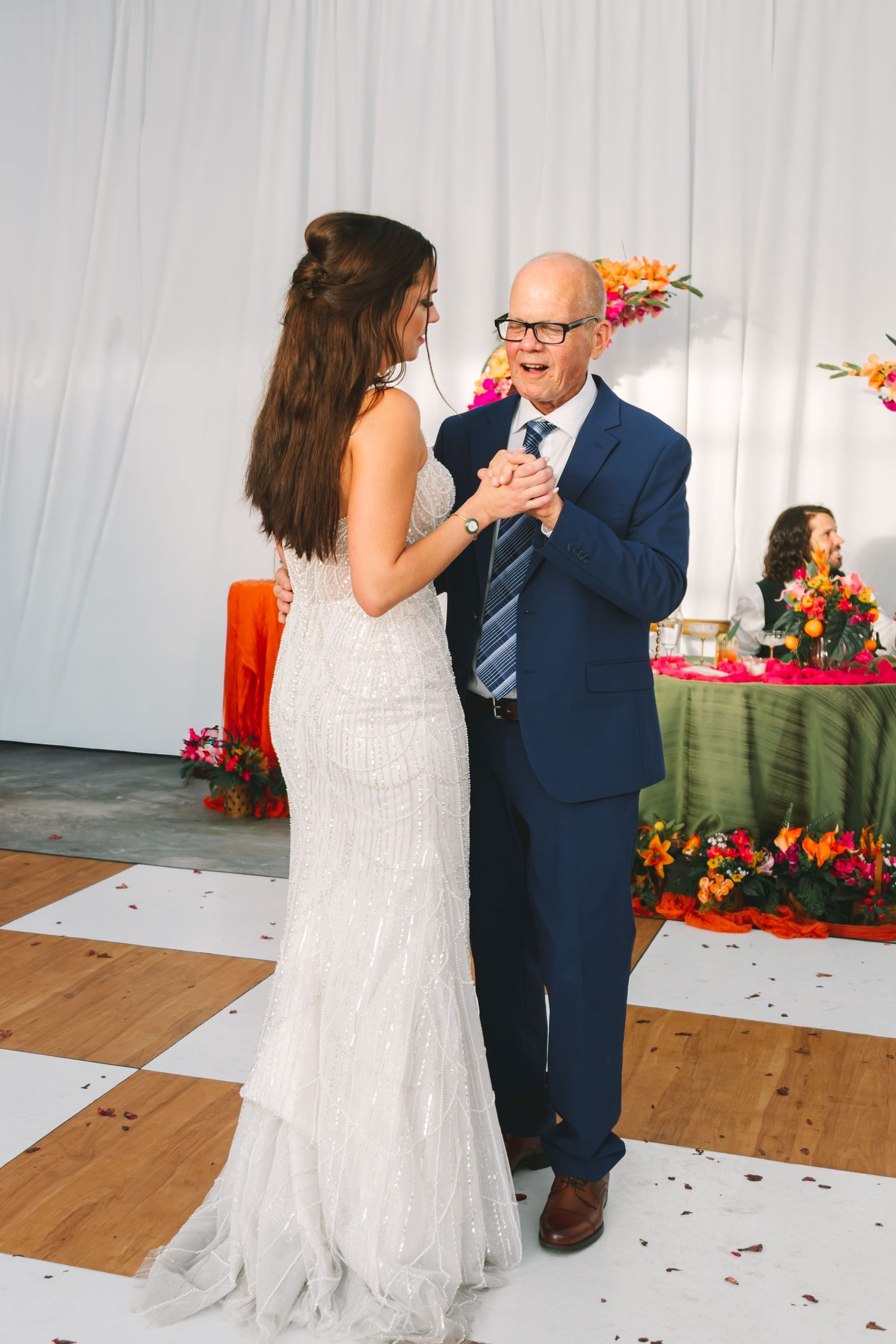 Bride in white lace gown shares father-daughter dance with older man in navy suit at wedding reception.