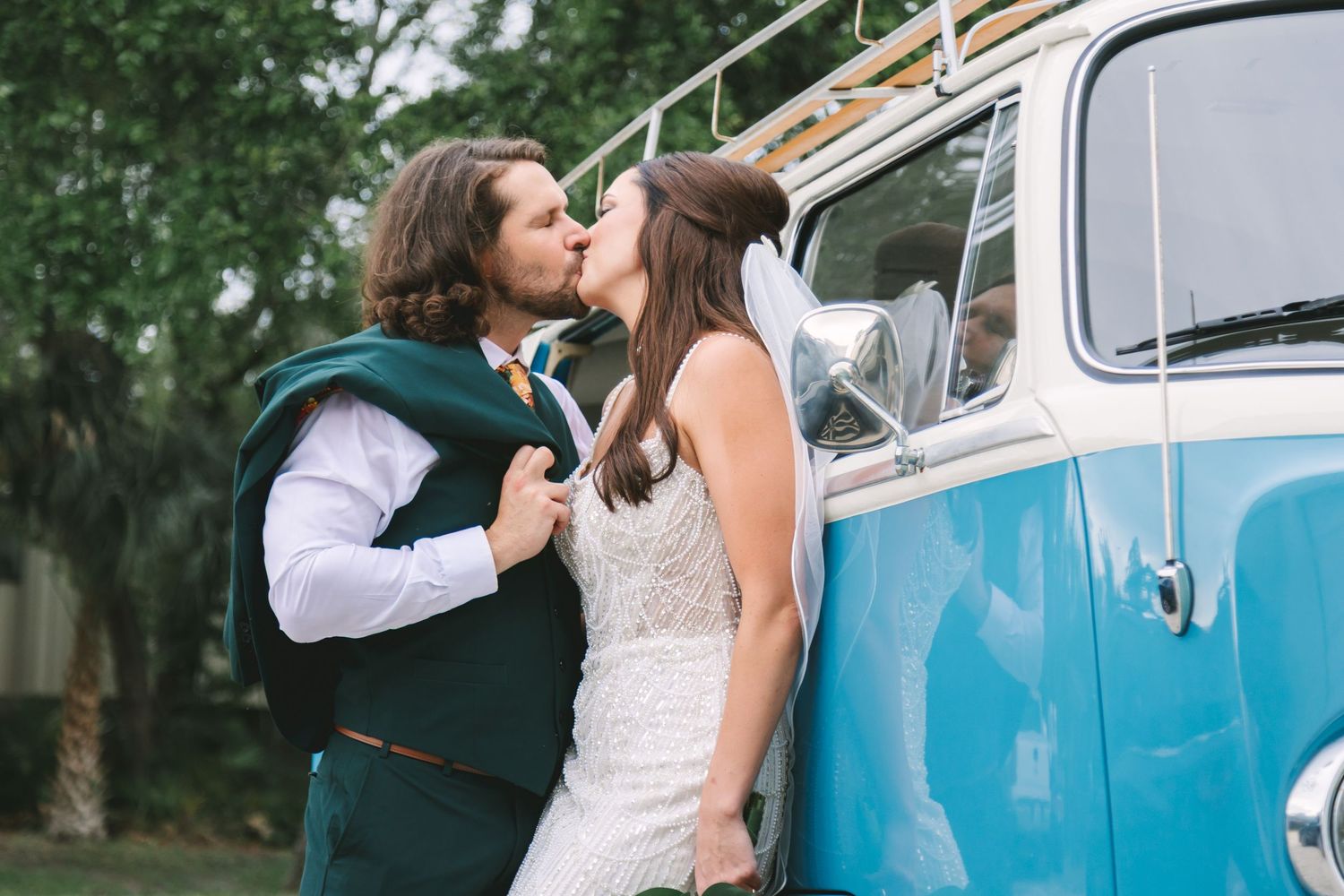 Bride and groom share a kiss beside a vintage blue VW bus on their wedding day.