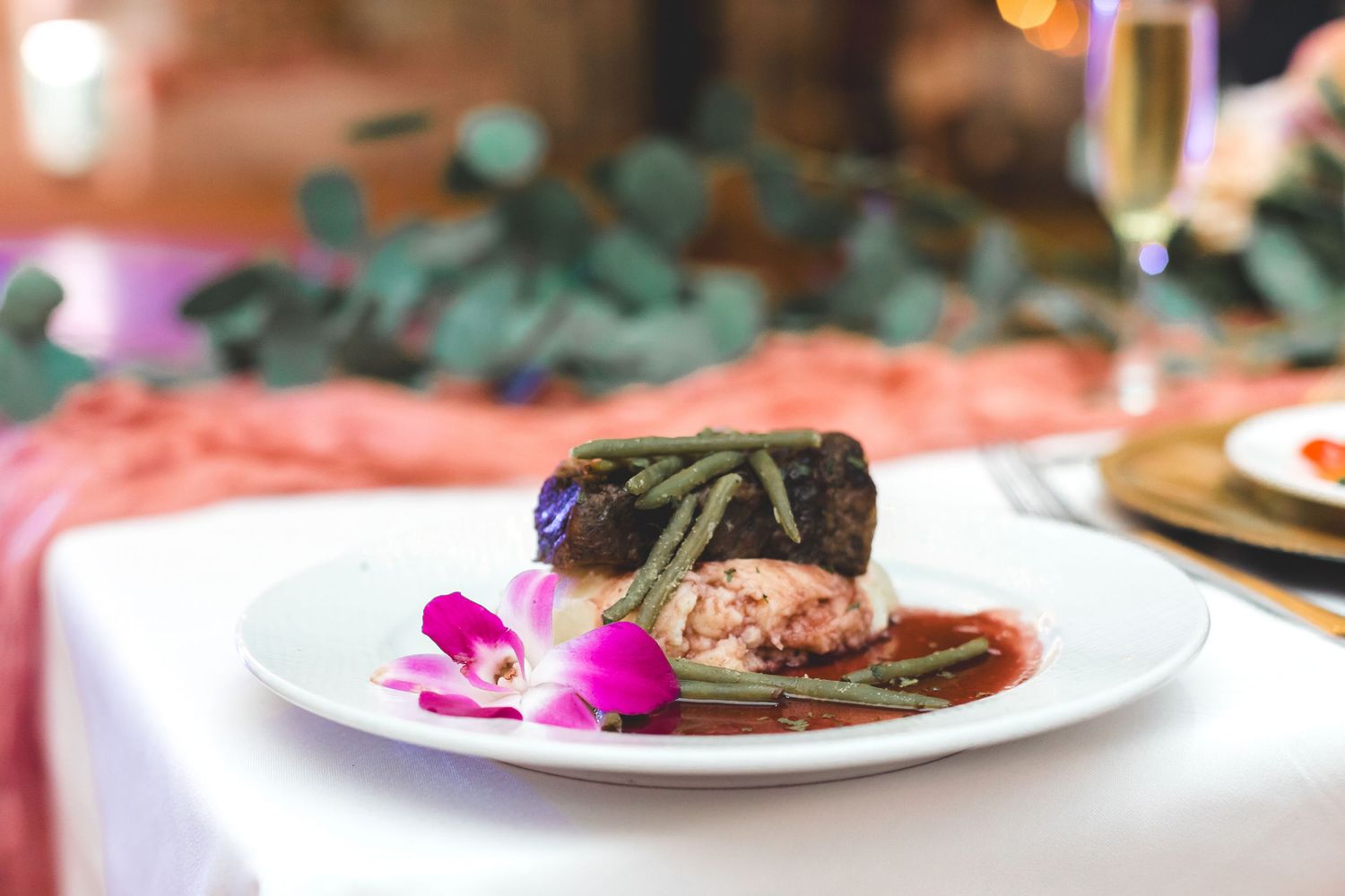 Elegant plated wedding dinner with filet mignon, green beans, and pink orchid garnish on a white tablecloth.