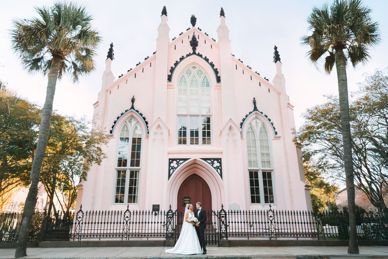Bride and groom pose before a stunning pink Gothic Revival church flanked by palm trees in Charleston, SC.
