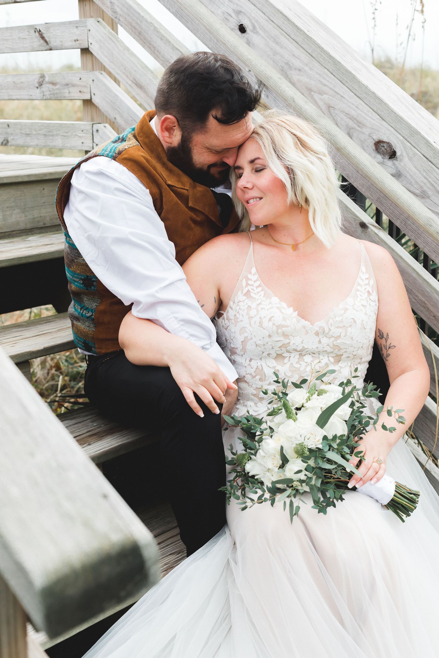 Couple embracing on wooden beach stairs, bride holding white bouquet in lace wedding gown.
