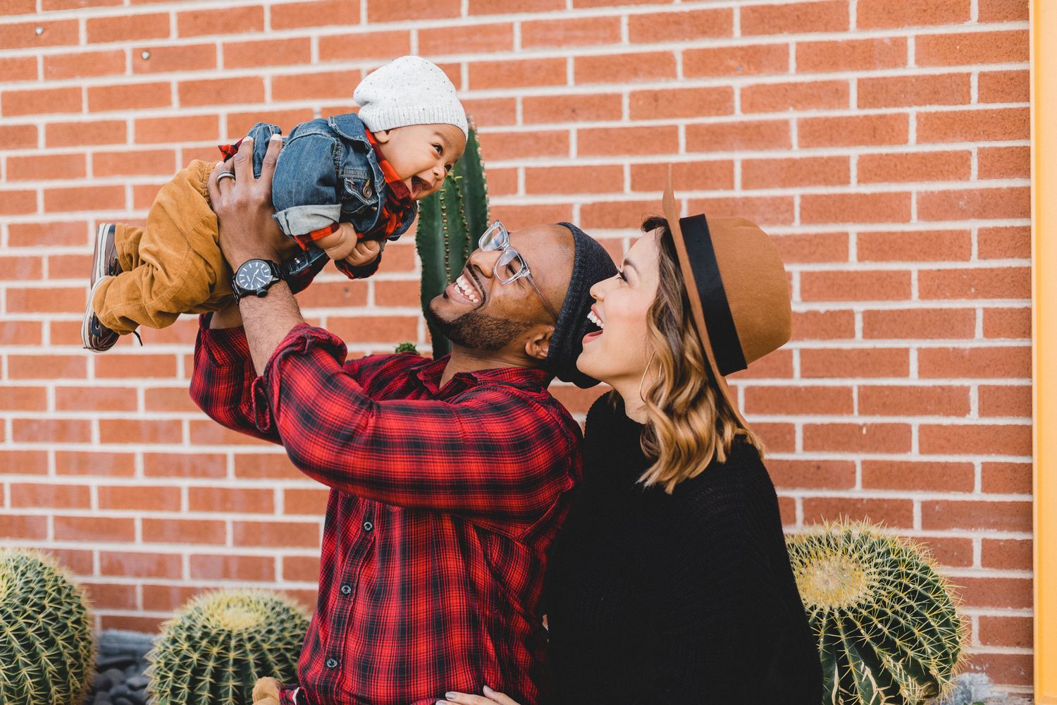 Joyful couple lifting smiling baby in front of brick wall with cacti in warm sunlight.