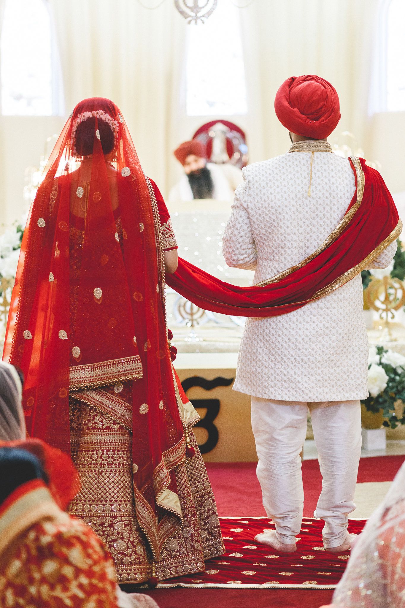 Sikh bride in red lehenga and groom in white sherwani walking during Anand Karaj wedding ceremony.