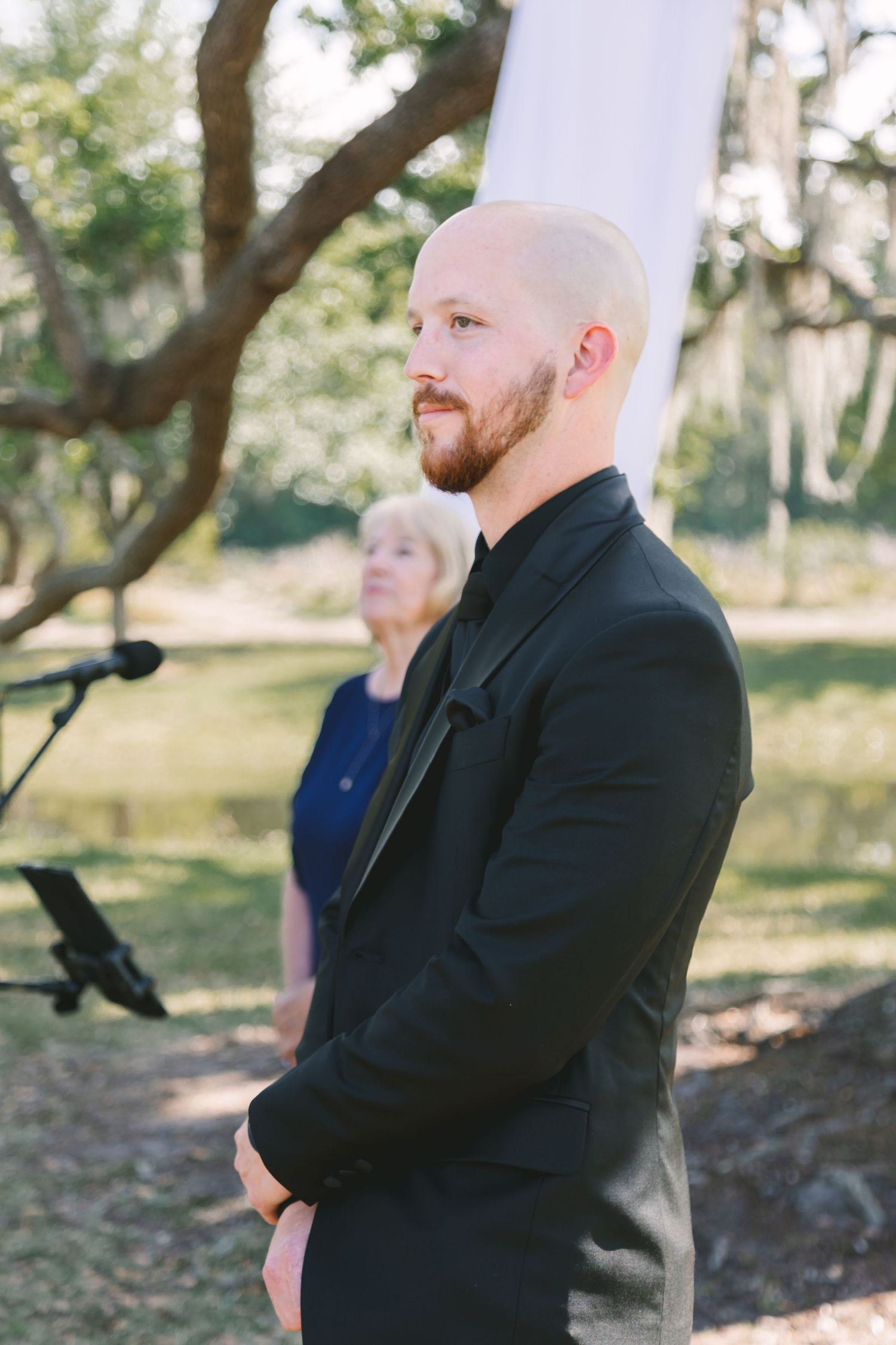 Bald groom with beard in black suit standing at outdoor wedding ceremony among trees.